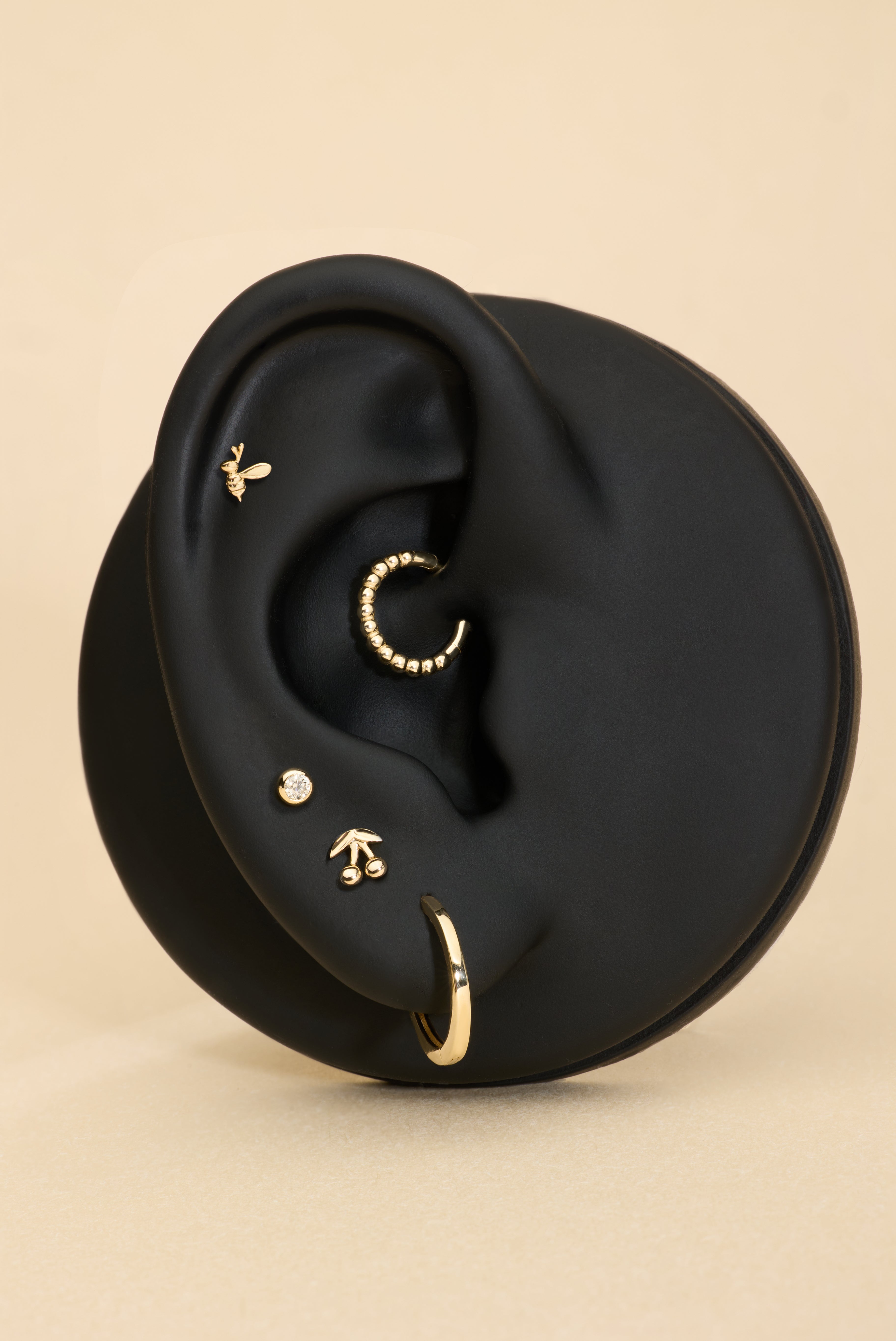 A black ear model displays five gold earrings, including the 14K Solid White Gold Bumble Bee Flat Back Stud (Left), with a beige background.