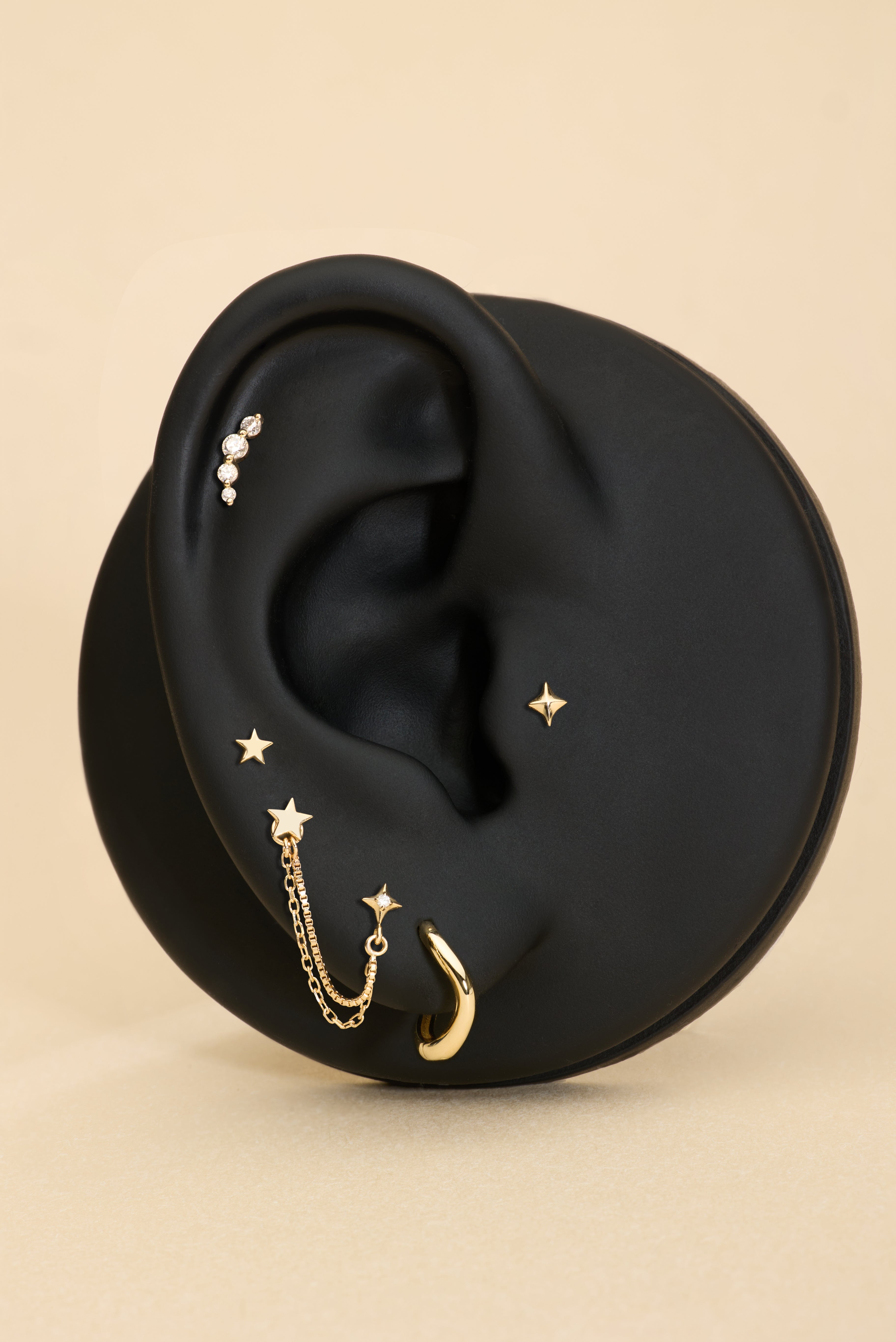 A black ear-shaped display showcases the 14K Solid Yellow Gold Four Point Star Flat Back Stud with other jewelry pieces against a beige background.
