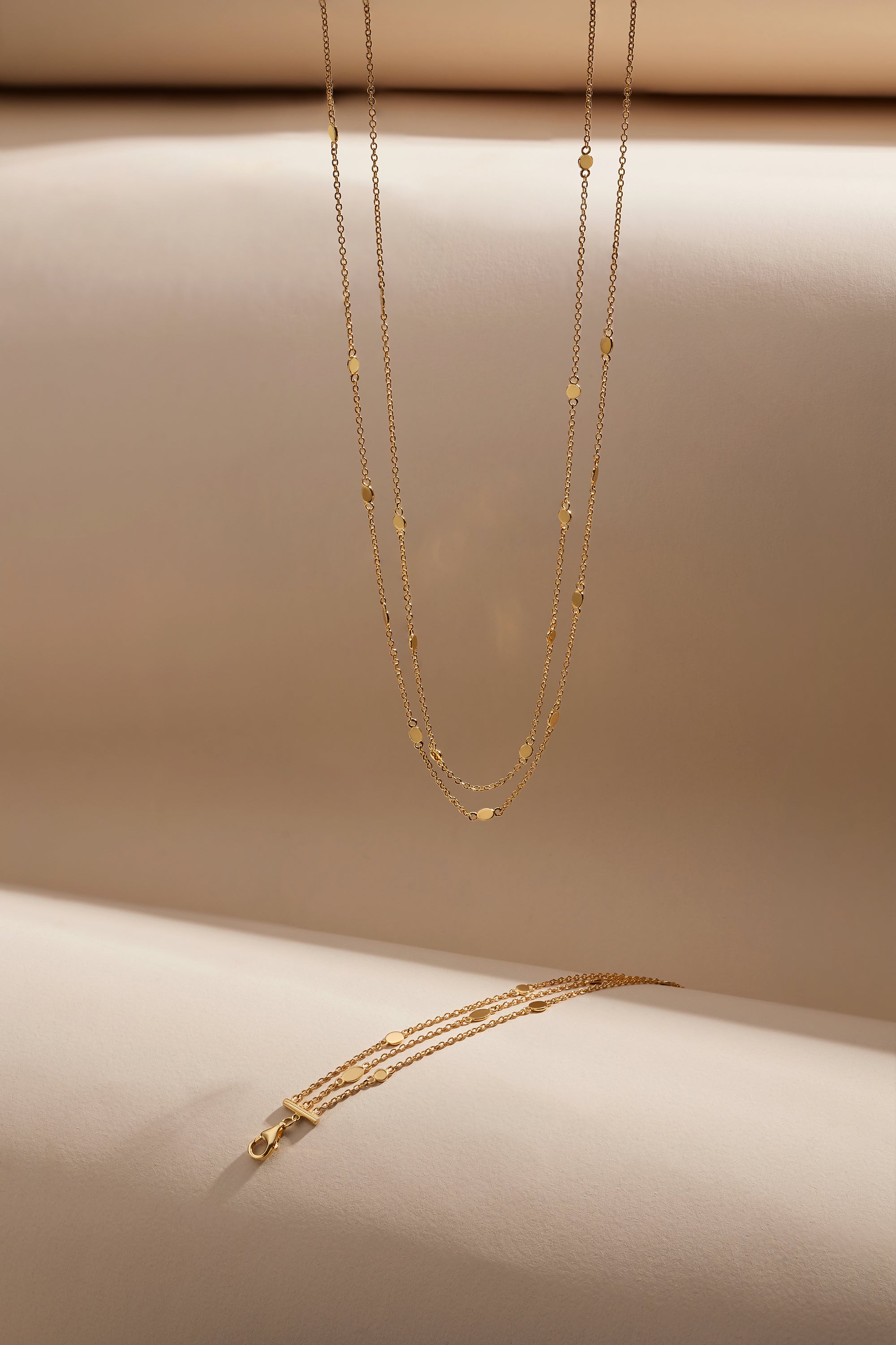 The Simona Double Chain Stacking Necklace in Gold features delicate double chains with small bead accents, displayed vertically and horizontally against a soft beige background.