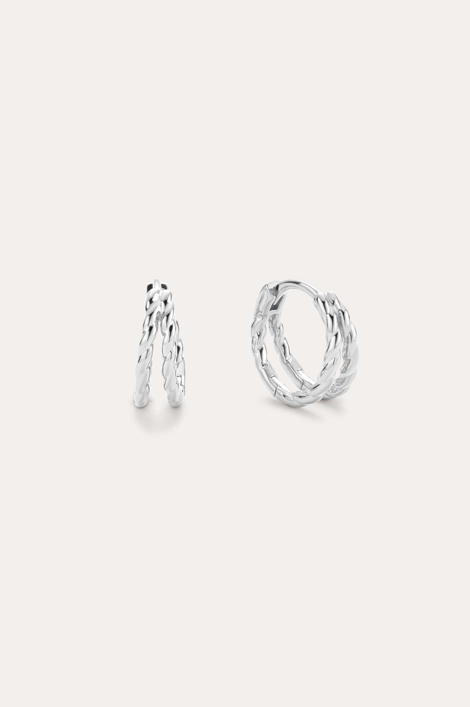 The Mae Twist Double Huggie Earrings in Silver feature a twisted, leafy design. Displayed on a plain background, one earring stands slightly open while the other lies flat, showcasing their intricate detailing.