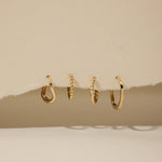 Four 14K recycled solid gold twisted huggie earrings are displayed in a row, hanging through a torn section of beige paper with a neutral background.