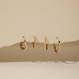 Four 14K recycled solid gold twisted huggie earrings are displayed in a row, hanging through a torn section of beige paper with a neutral background.