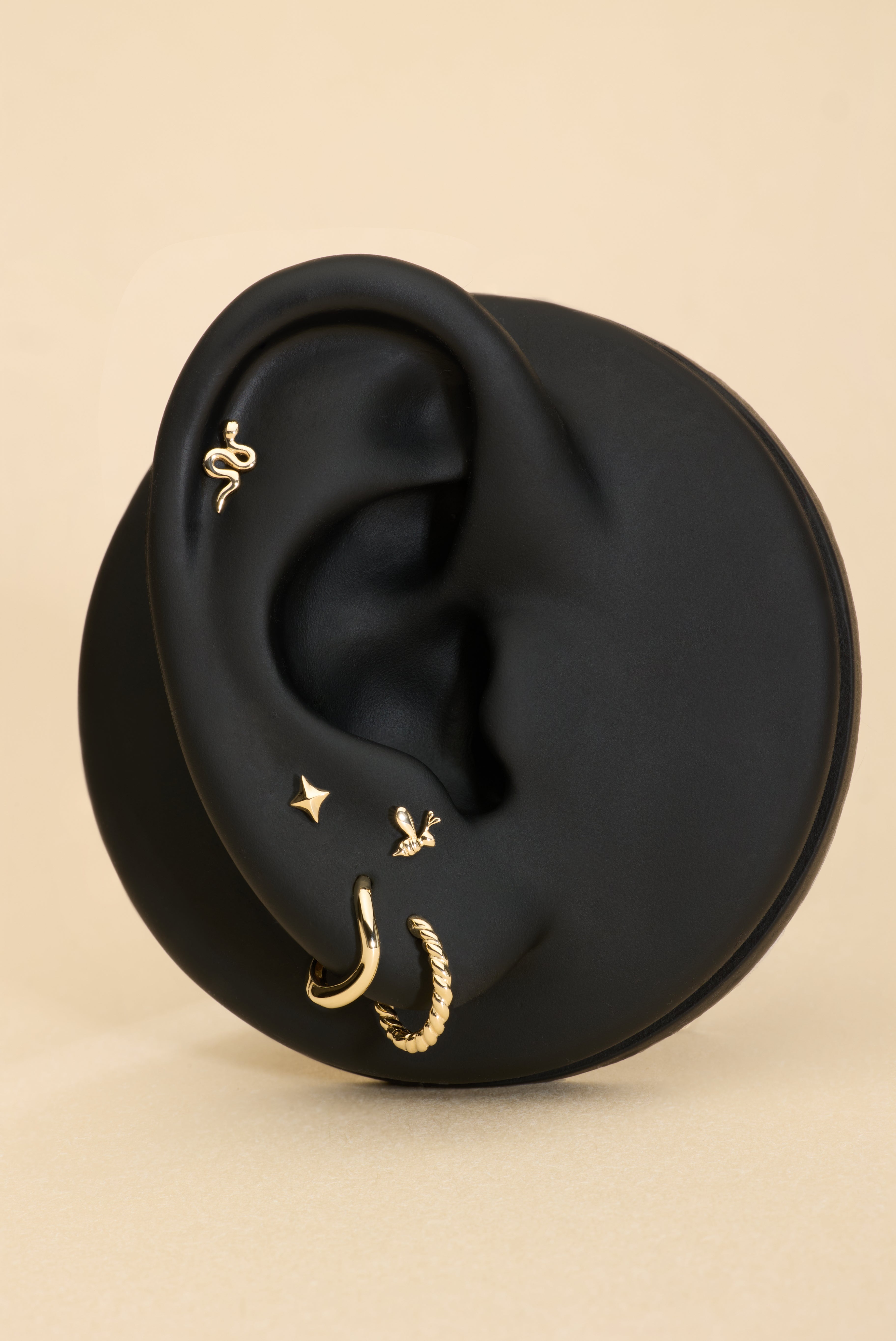 A black mannequin ear displays five gold earrings, including a 14K Solid Yellow Gold Snake Flat Back Stud at the top, alongside a small gold hoop, textured hoop, star stud, and bird earring, all set against a beige background.