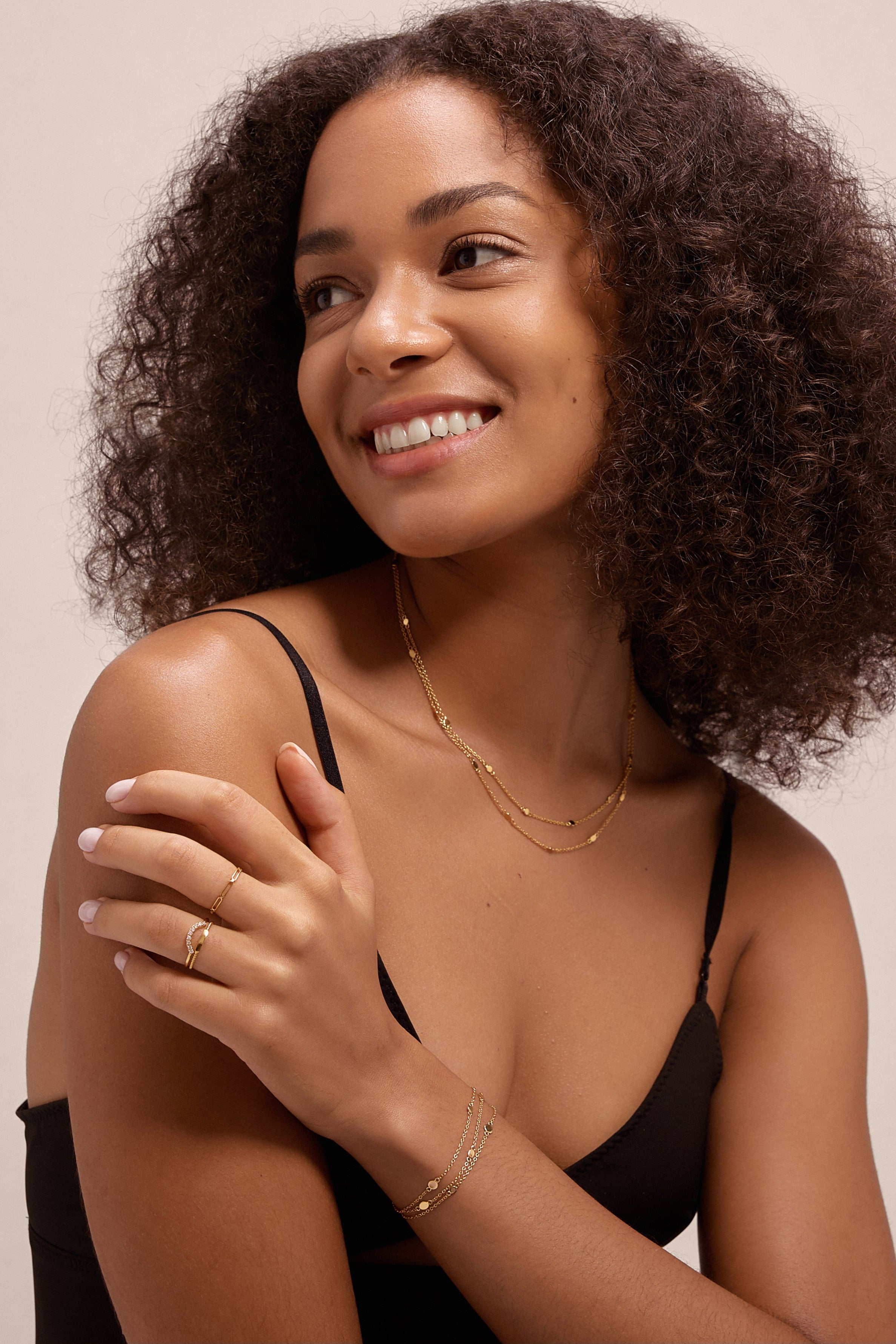 A woman with curly hair in a black tank top smiles to the side, showcasing the Simona Double Chain Stacking Necklace in Gold along with a gold bracelet and rings, set against a plain light background.
