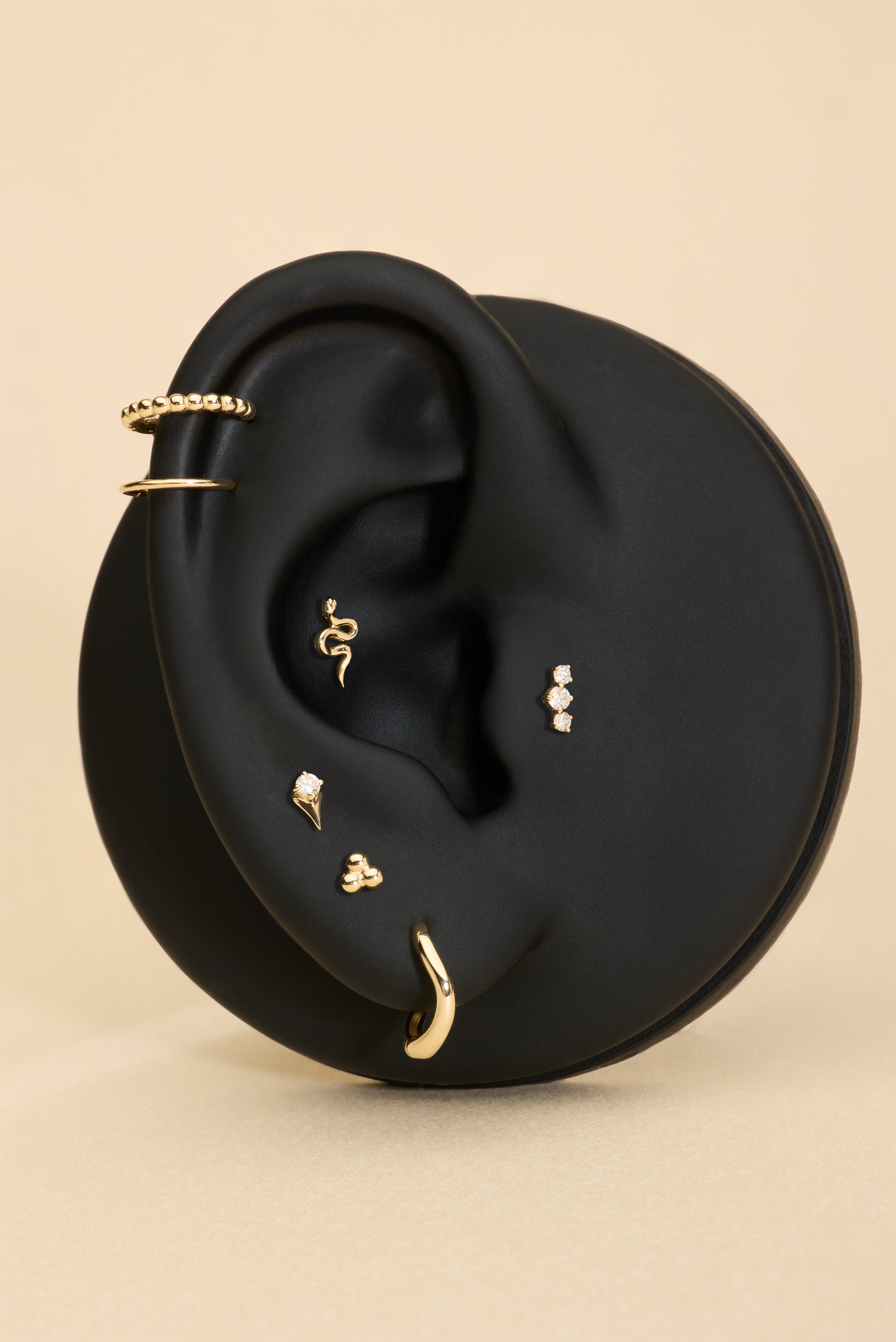 A black ear-shaped display showcases the 14K Solid Yellow Gold Snake Flat Back Stud among other gold earrings and studs—ideal for helix piercings—set against a beige background.