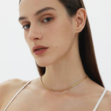 A woman with straight brown hair and natural makeup wears the Estelle Herringbone Chain Necklace in Gold with a white spaghetti strap top, posing against a plain light background.