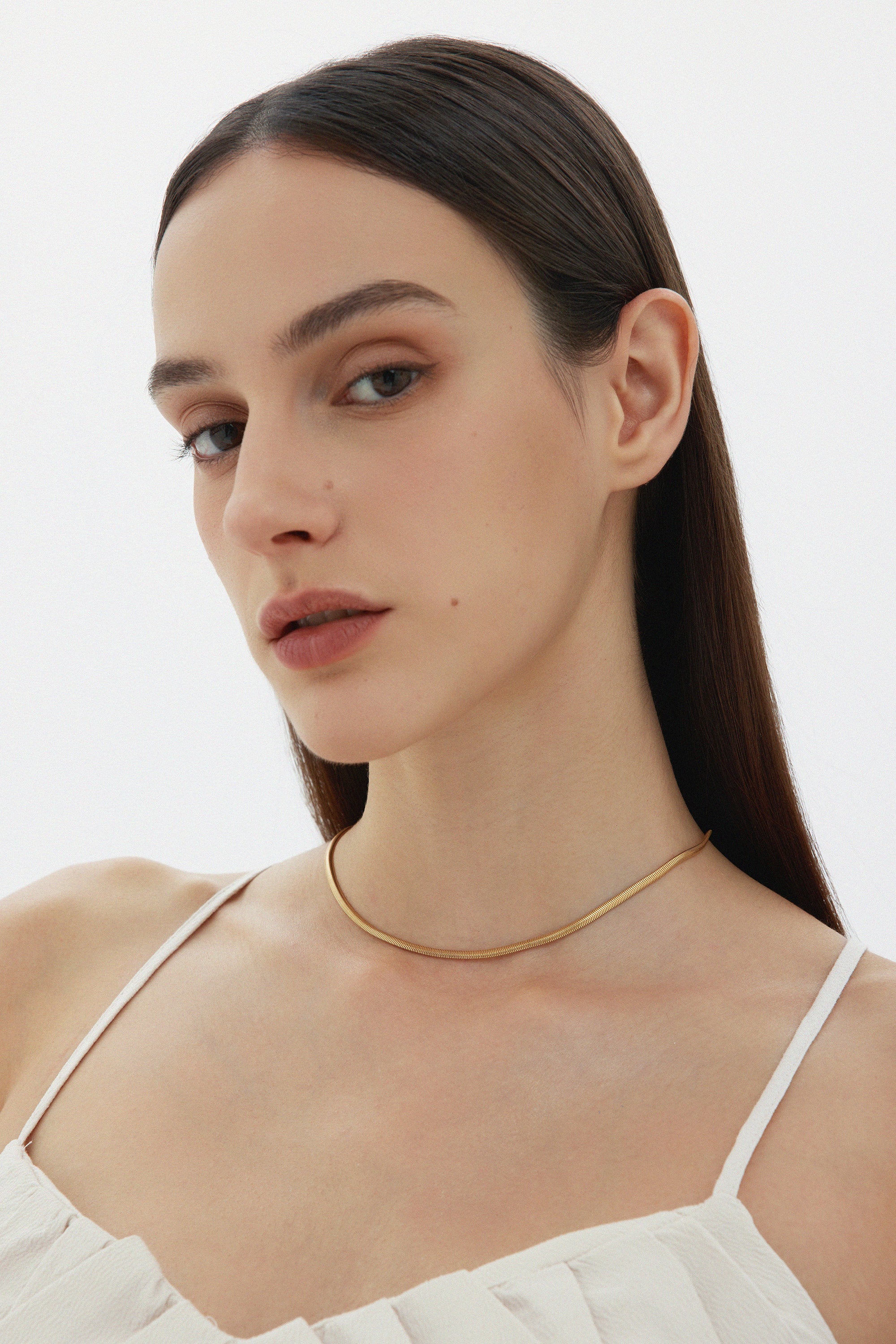 A woman with straight brown hair and natural makeup wears the Estelle Herringbone Chain Necklace in Gold with a white spaghetti strap top, posing against a plain light background.