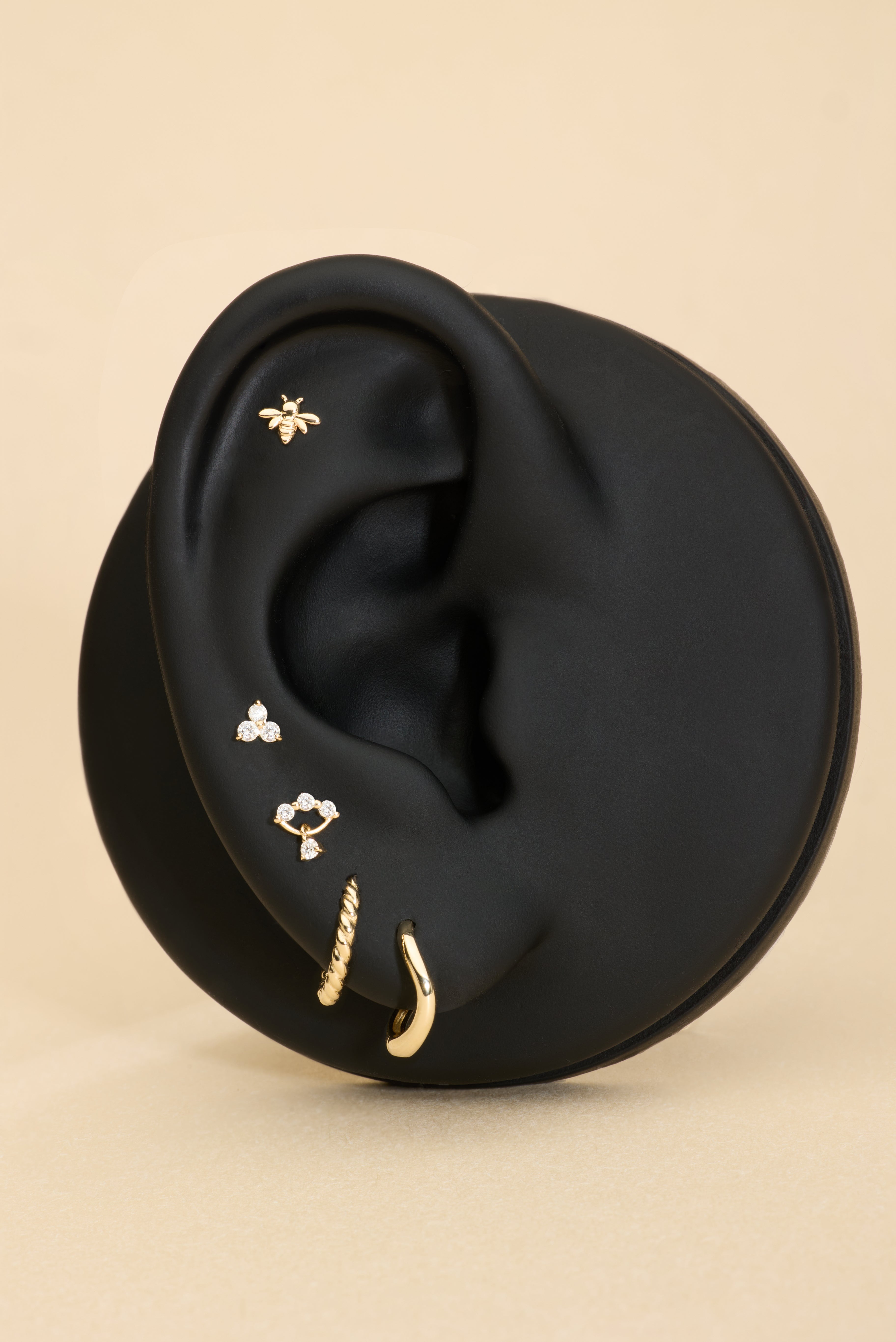 Black ear with gold huggie earrings, bee flatback stud, and diamond flatback stud on a beige background