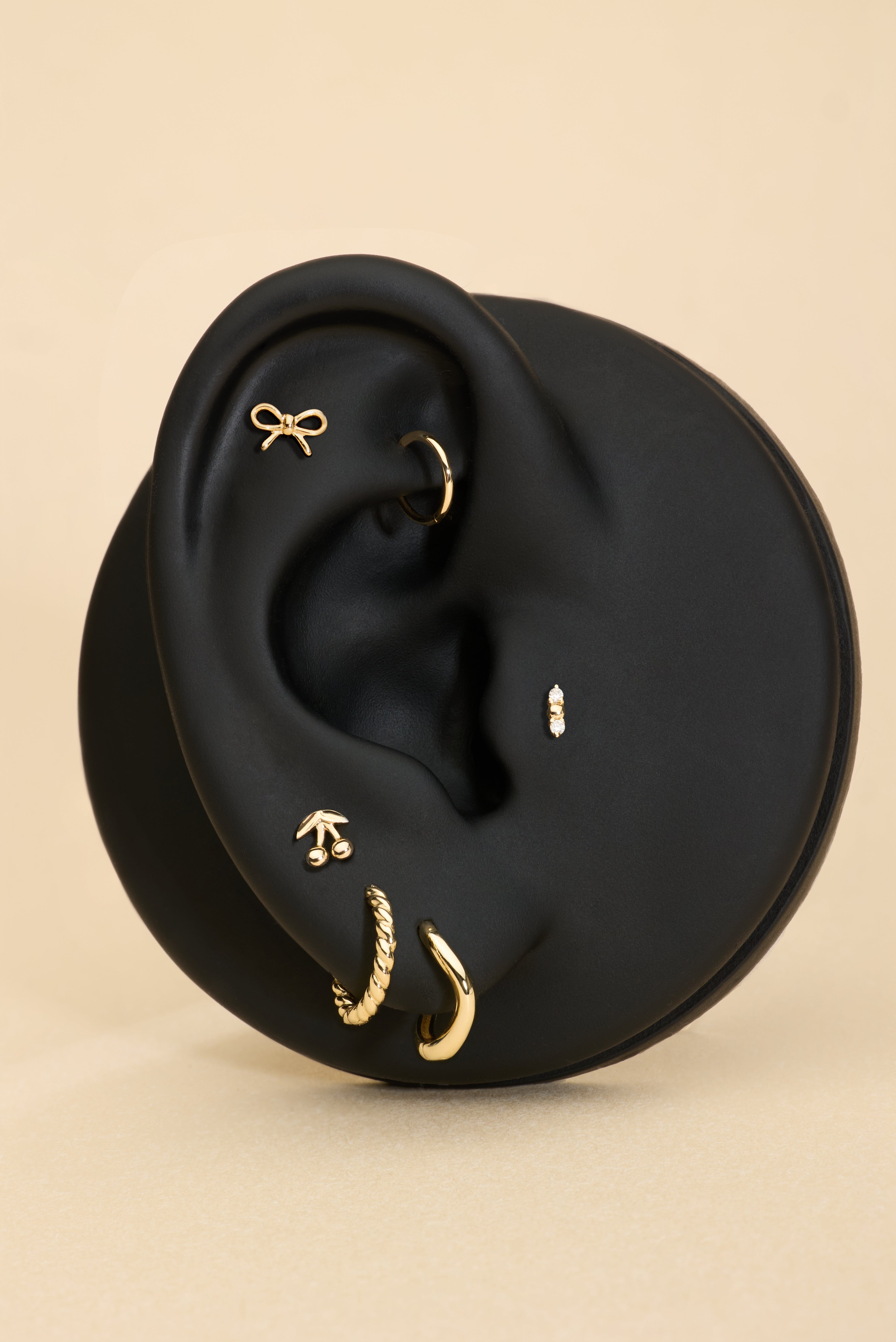 Ear with gold earrings, bow stud, cheery stud and diamond stud on a beige background