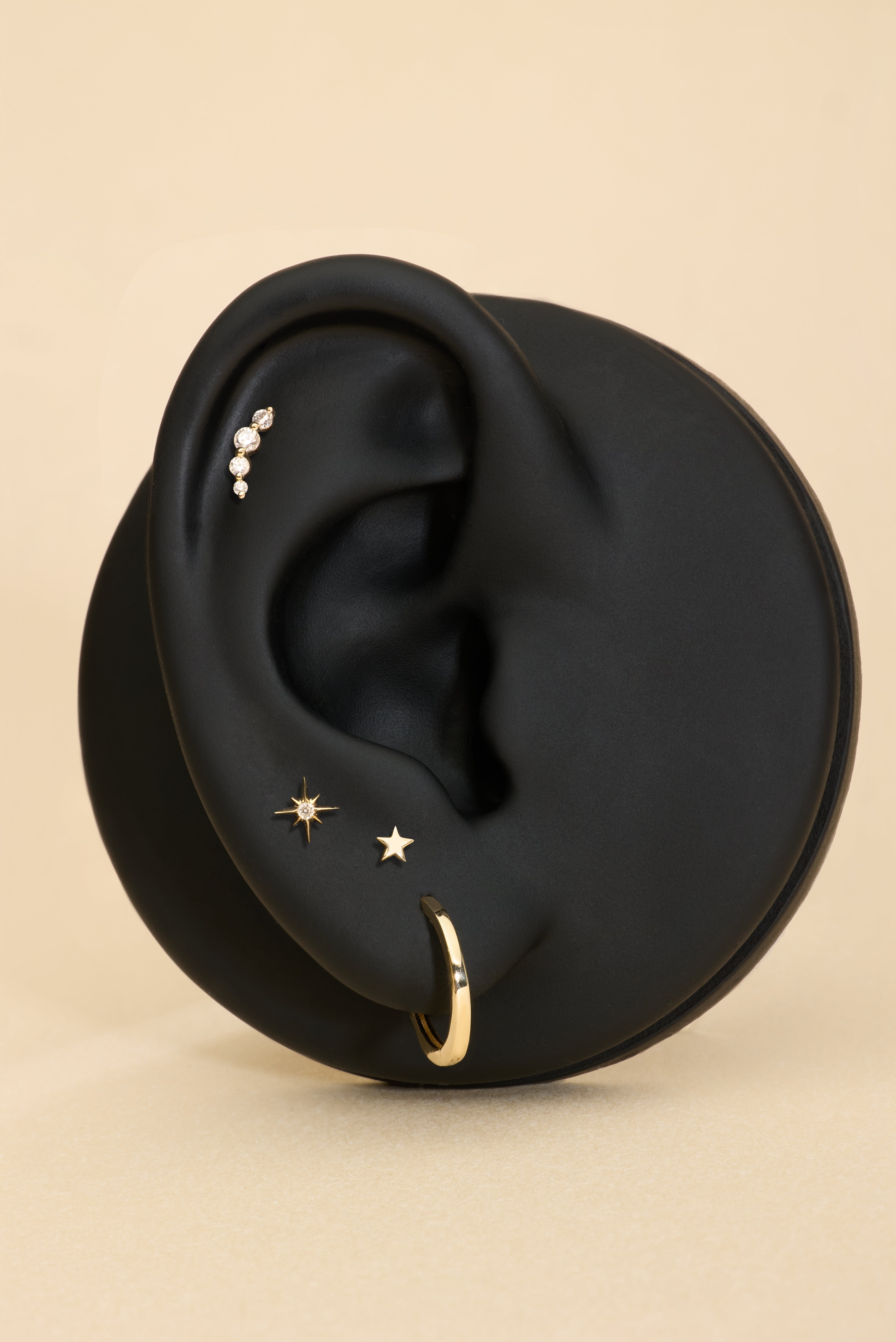 A black mannequin ear showcases the 14K Recycled Solid Yellow Gold Plain Huggie Earring and three other sustainable pieces, all set against a beige background.