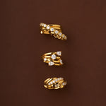 The Vina Crystal Bezel Ear Cuff in Gold, featuring sparkling cubic zirconia and both smooth and textured gold finishes, is elegantly displayed on a brown background.