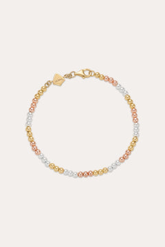 The Cecelia Three Tone Trinity Bracelet features small gold, sterling silver, and rose gold beads in a gradient pattern with a gold clasp and tag. This dainty bracelet is 18k gold plated and styled on a light background.