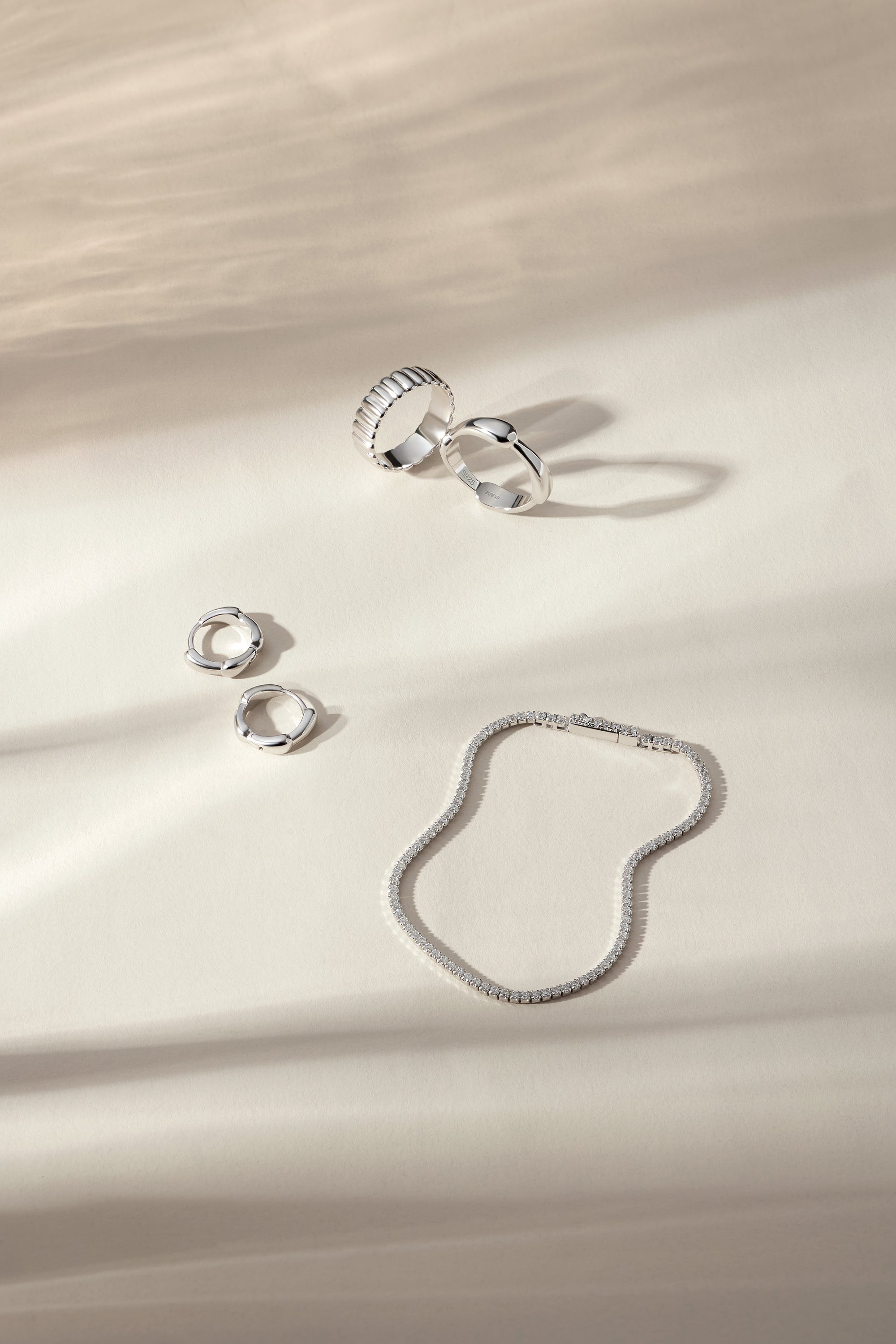 Four silver rings, including the Charlotte Eternity Ridged Ring in Silver, and a silver chain necklace are displayed on a cream-colored surface with soft shadows from gentle lighting.