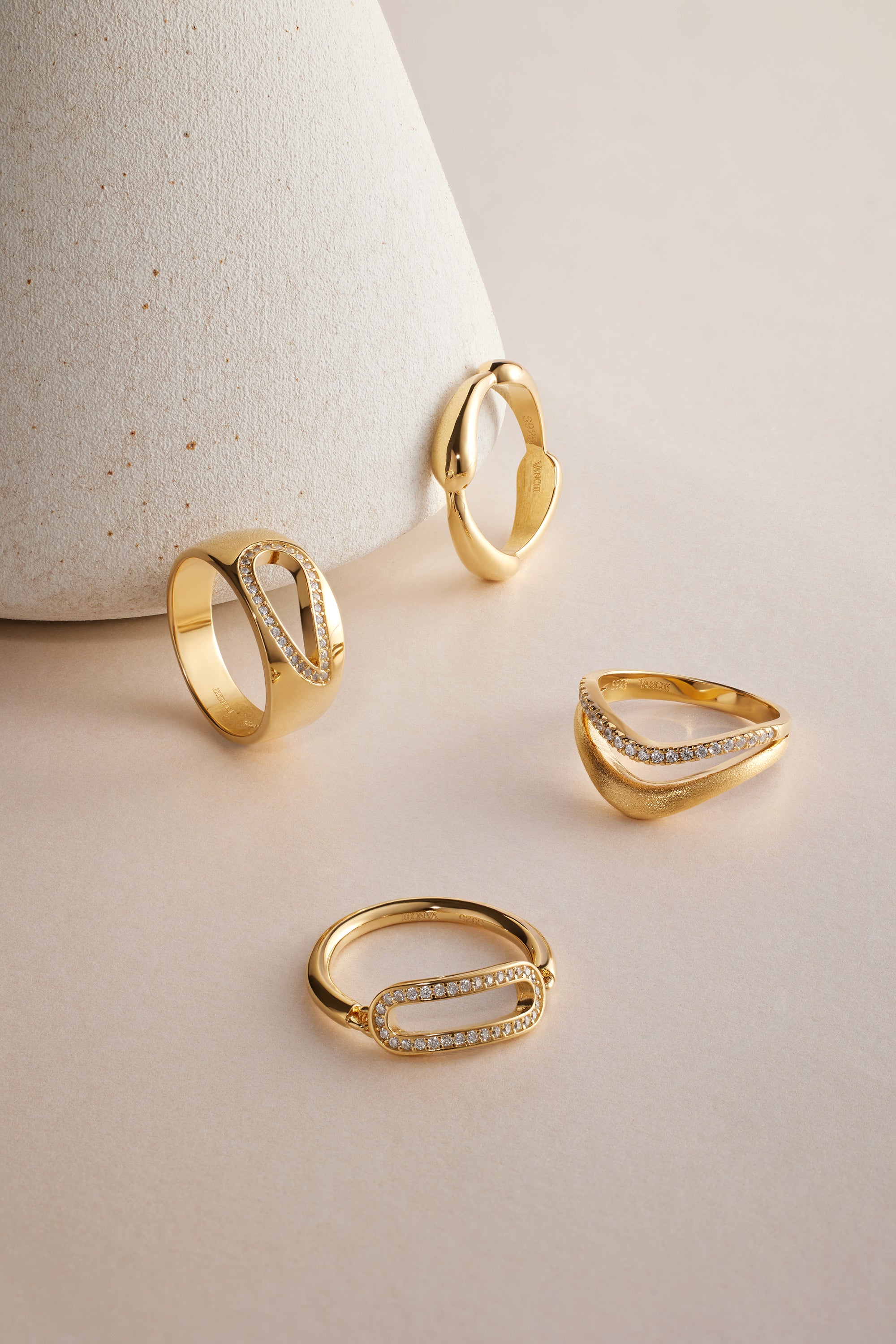 Four gold rings, including the striking Colette Link Droop Ring in Gold with its geometric design and small diamonds, are displayed on a pale surface, with one ring leaning against a textured, cone-shaped object.