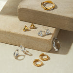 A display of gold and silver jewelry, featuring rings, hoop earrings, and the Colette Link Droop Ring in Gold, arranged on textured beige platforms under soft lighting.