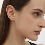 A woman with straight brown hair wears the Vina Crystal Bezel Ear Cuff in Gold, featuring cubic zirconia on her upper ear. She sports natural makeup and a black top, set against a plain background.