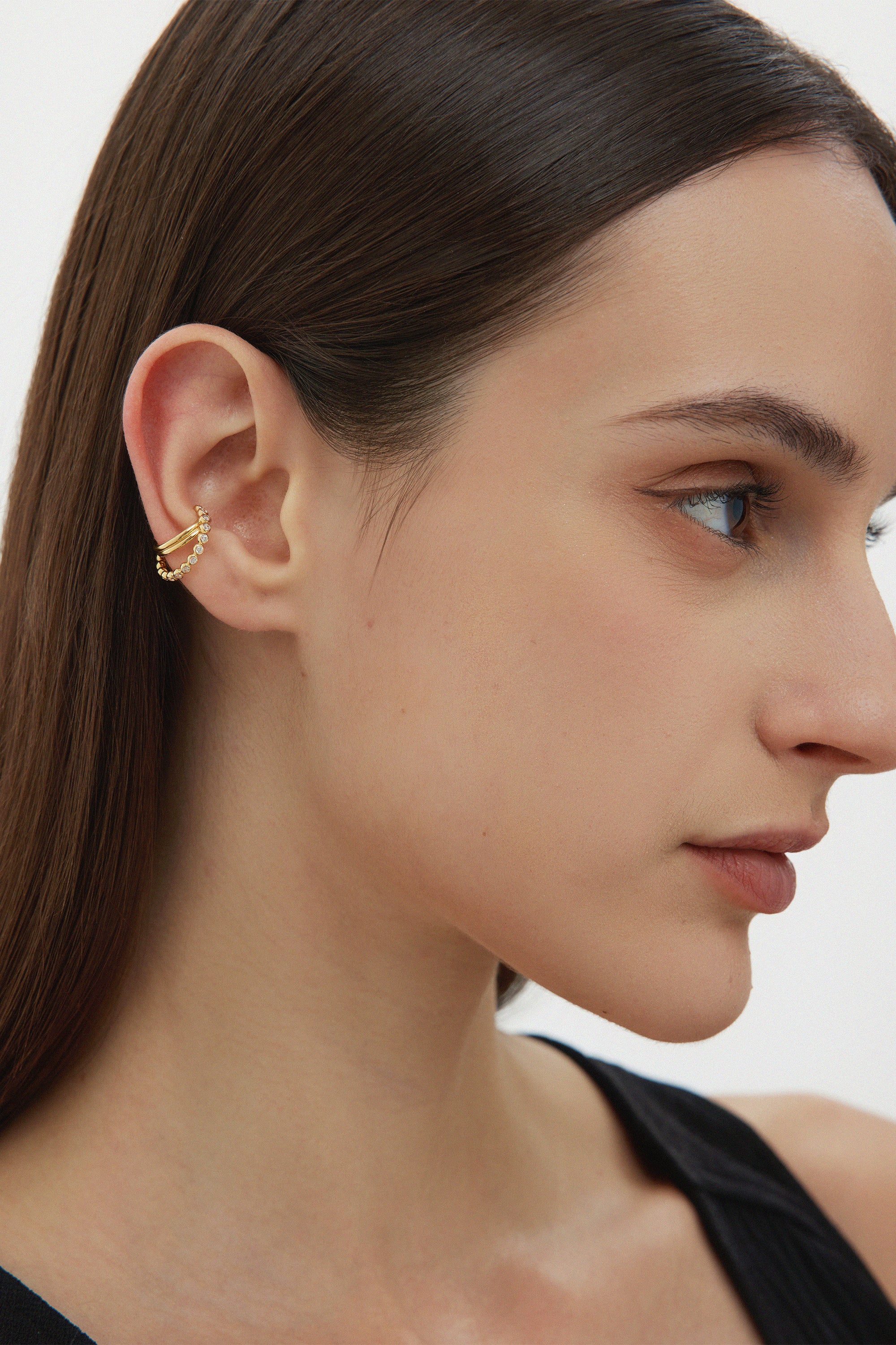 A woman with straight brown hair wears the Vina Crystal Bezel Ear Cuff in Gold, featuring cubic zirconia on her upper ear. She sports natural makeup and a black top, set against a plain background.
