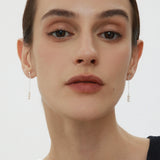 A person with fair skin and dark hair pulled back is wearing subtle makeup and the Eden Trio Pearl Drop Earrings in Silver. They are dressed in a sleeveless black top and looking directly at the camera against a plain background.