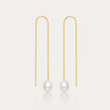 The Eira Pearl Drop Threader Earrings in Gold feature delicate chains ending with a single round white pearl, offering a minimalist and elegant look against a plain white background.