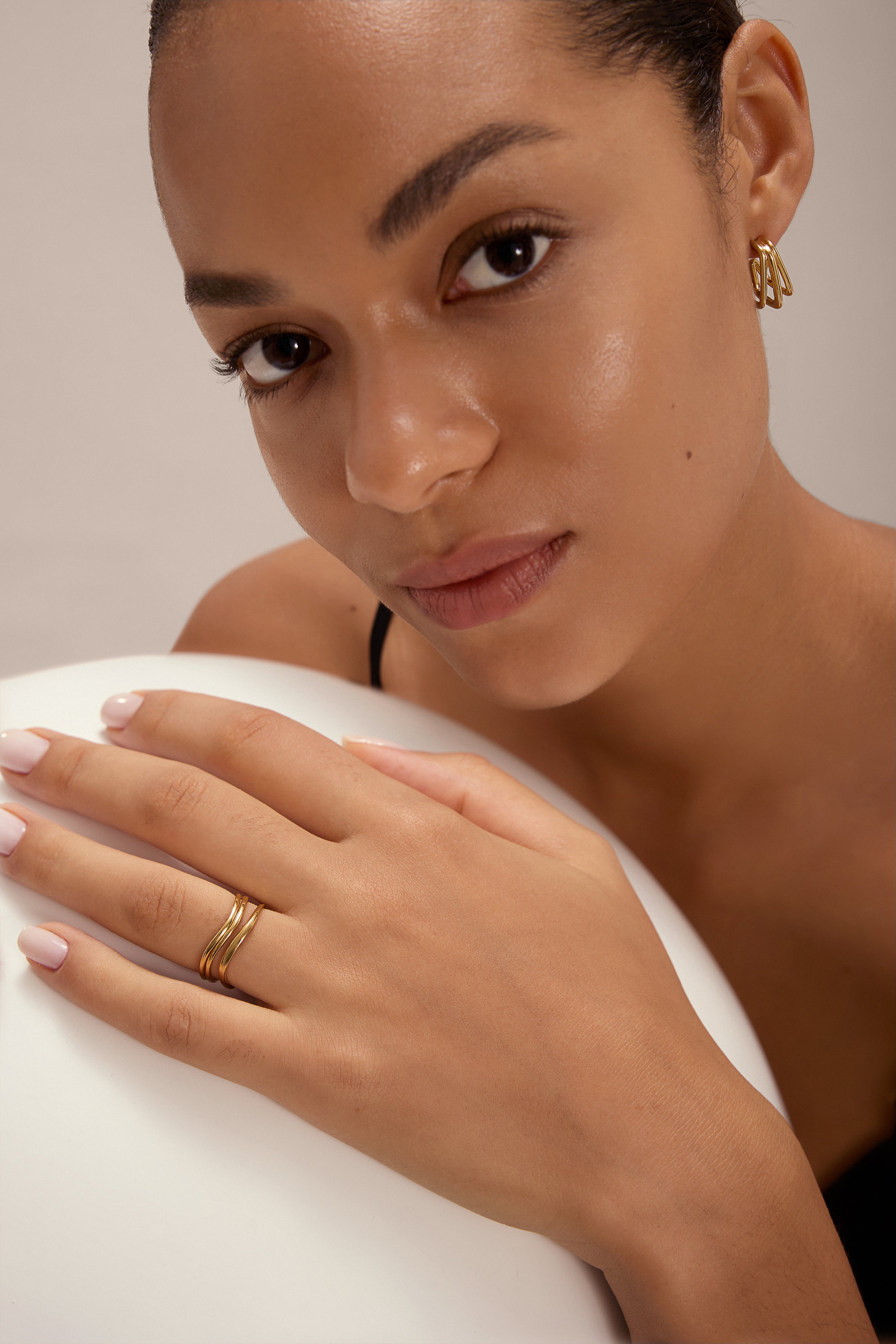 A woman with glowing skin and natural makeup wears Esther Square Hoop Stud Earrings in Gold. She rests her manicured hand on a white surface, gazing softly at the camera.