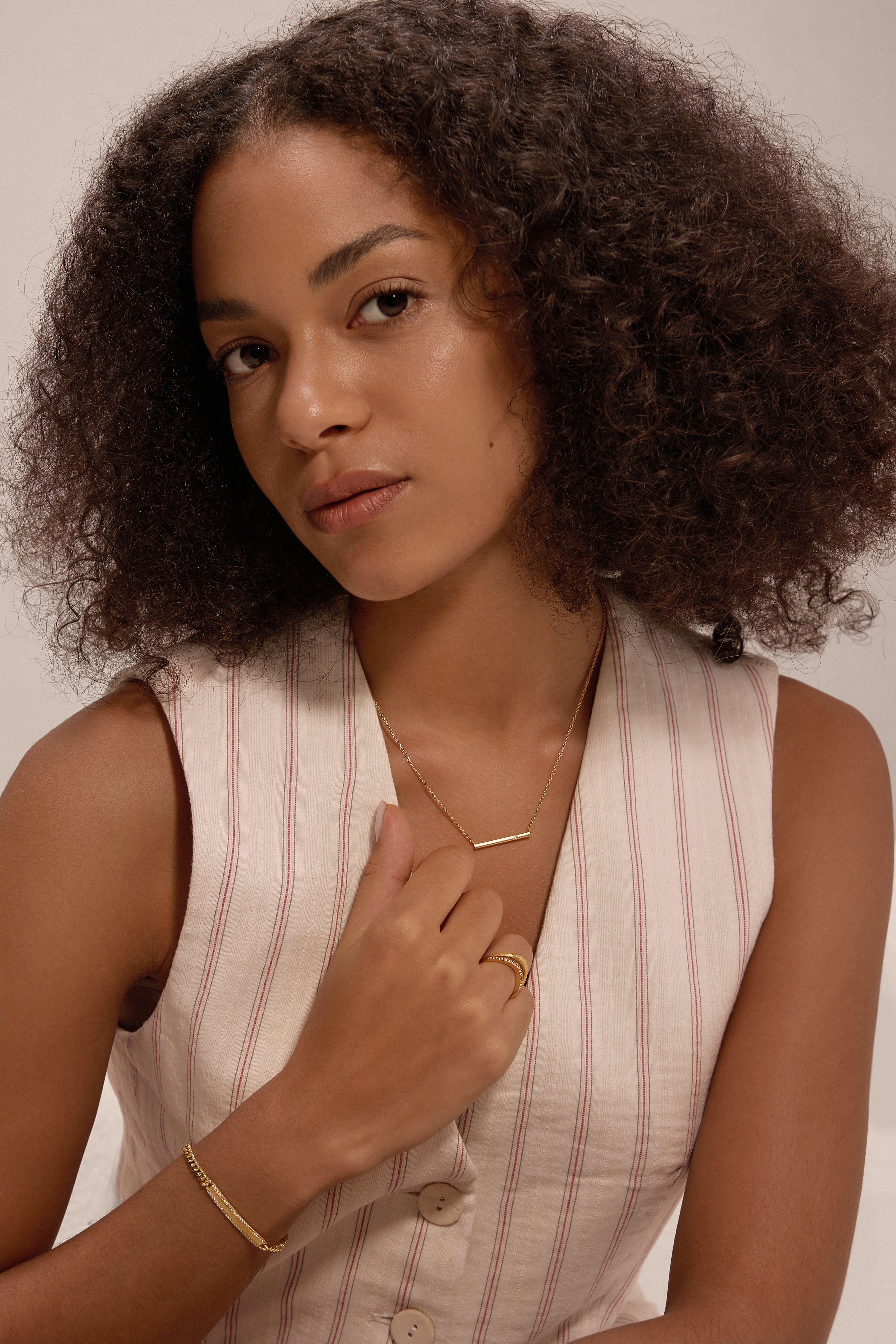 A woman with curly hair in a sleeveless, light-striped vest gazes softly at the camera. She wears gold jewelry, including a cubic zirconia ring and the Gisèle Cuban Link Bar Bracelet in Gold, and has a calm, neutral expression.