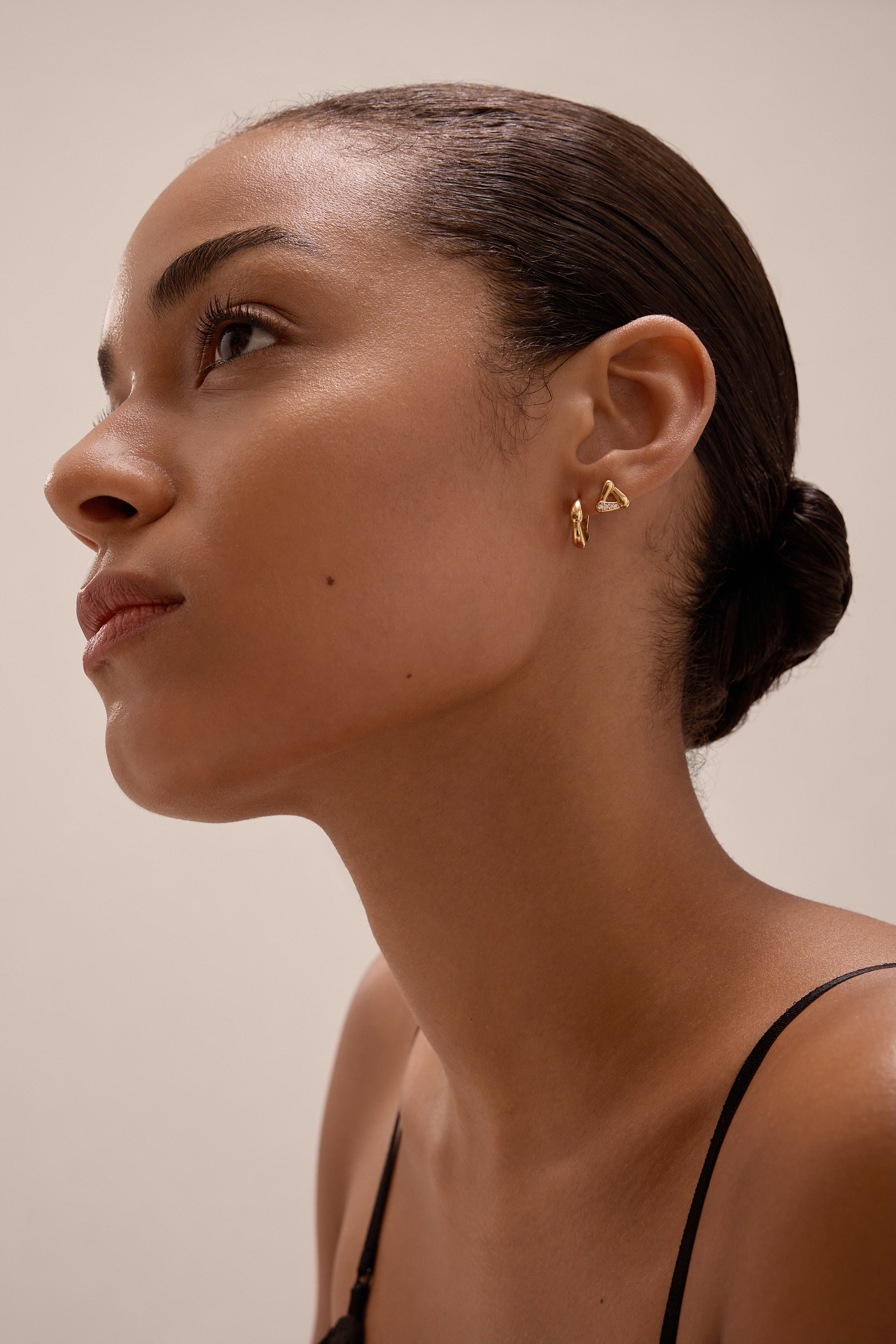 A woman with smooth, medium-brown skin and dark hair in a low bun gazes upward, wearing Colette Droop Link Studs in Gold and a black spaghetti strap top against a neutral background.