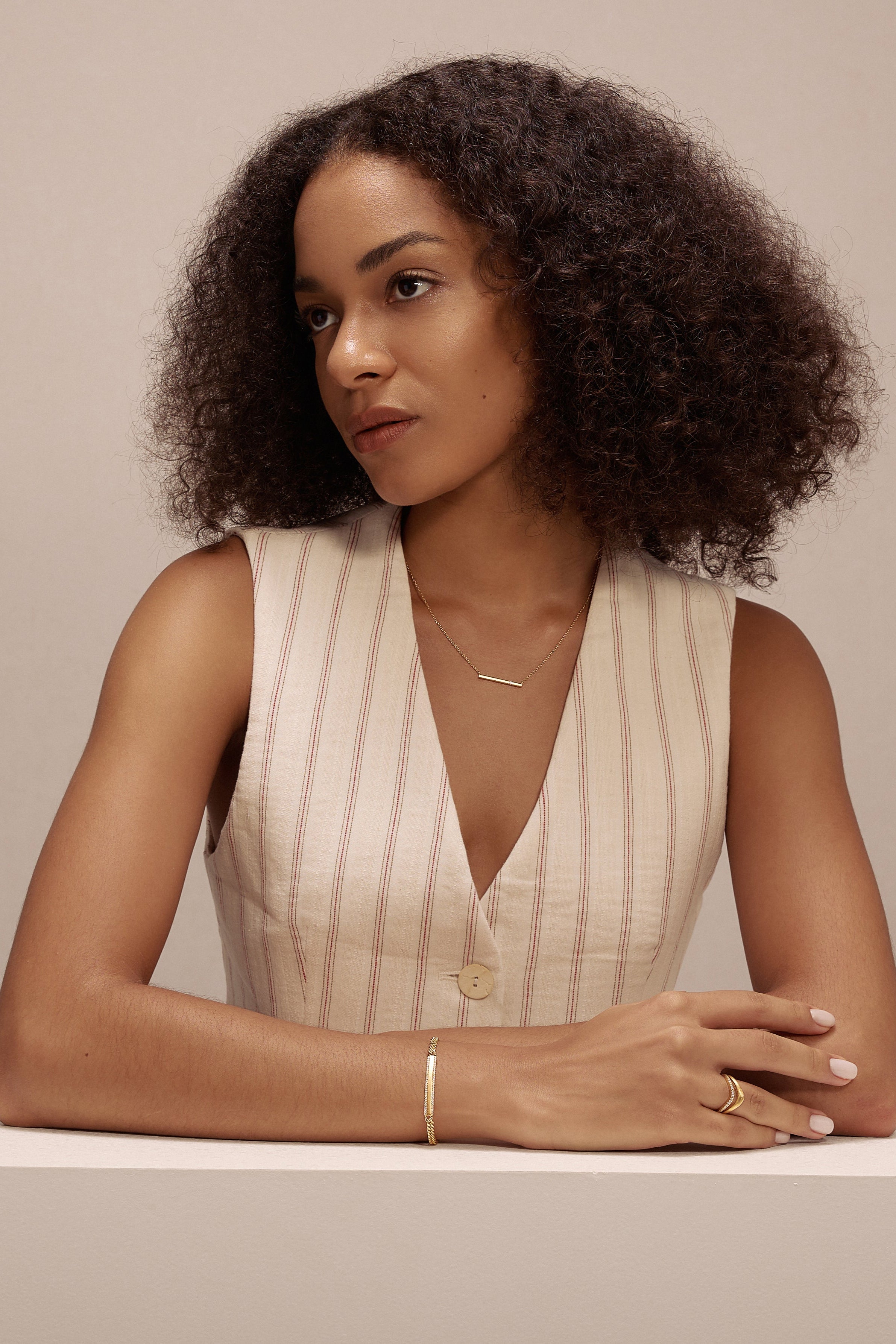 A woman with curly hair in a sleeveless, light top sits at a white table, looking to the side. She has a calm expression and wears delicate cubic zirconia jewelry and the Gisèle Cuban Link Bar Bracelet in Gold.