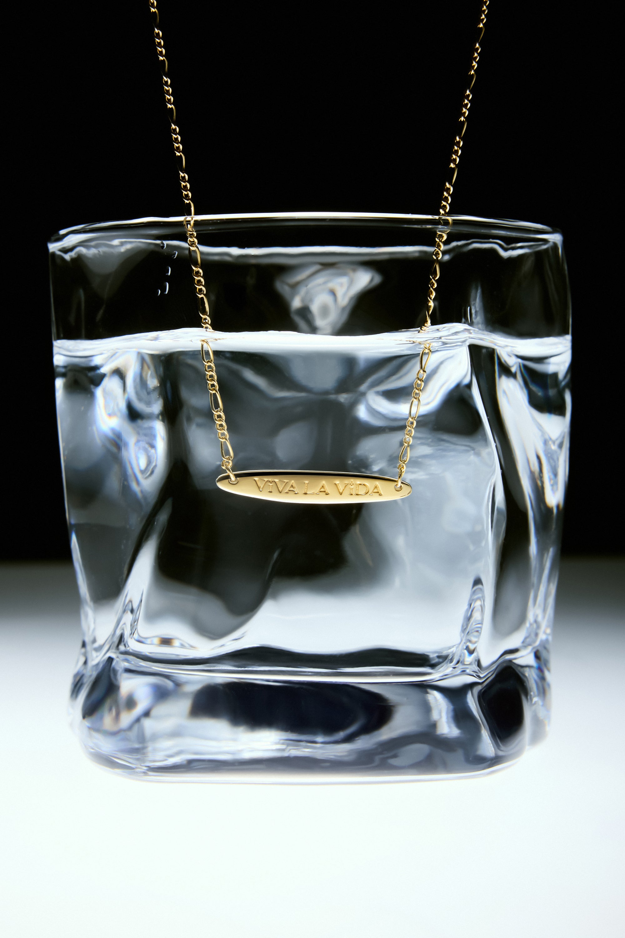 The Viva La Vida Bar Chain Necklace in Gold hangs over the rim of a clear glass filled with water, set against a white and black background, and features an adjustable figaro chain for the perfect fit.