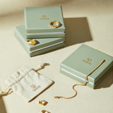 Three sage green VANCII jewelry boxes with gold jewelry—rings, a bracelet, and Judith Hexagon Angular Huggies in Gold—are displayed on a beige surface next to a cream drawstring pouch featuring the VANCII logo.