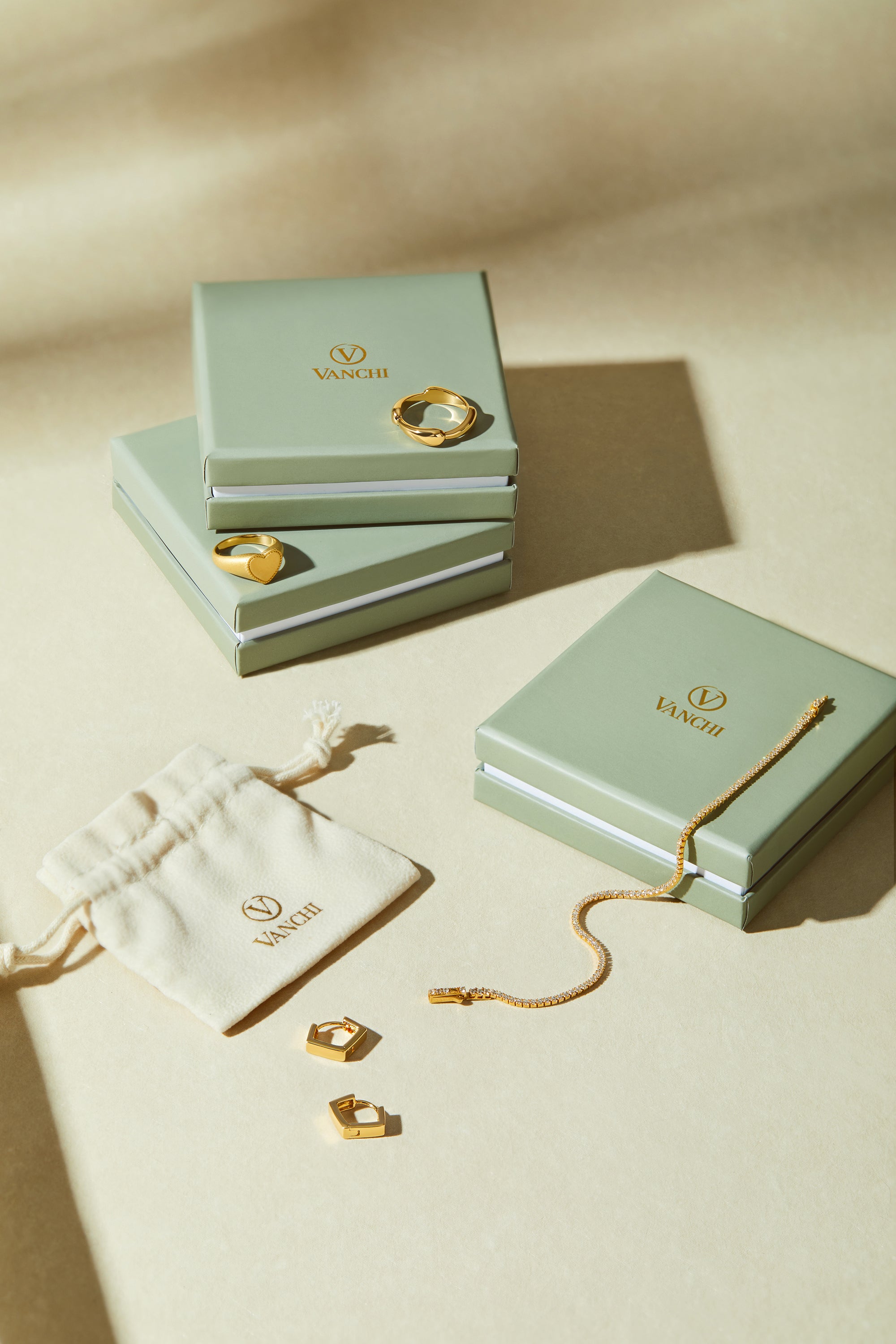 Three sage green VANCII jewelry boxes with gold jewelry—rings, a bracelet, and Judith Hexagon Angular Huggies in Gold—are displayed on a beige surface next to a cream drawstring pouch featuring the VANCII logo.