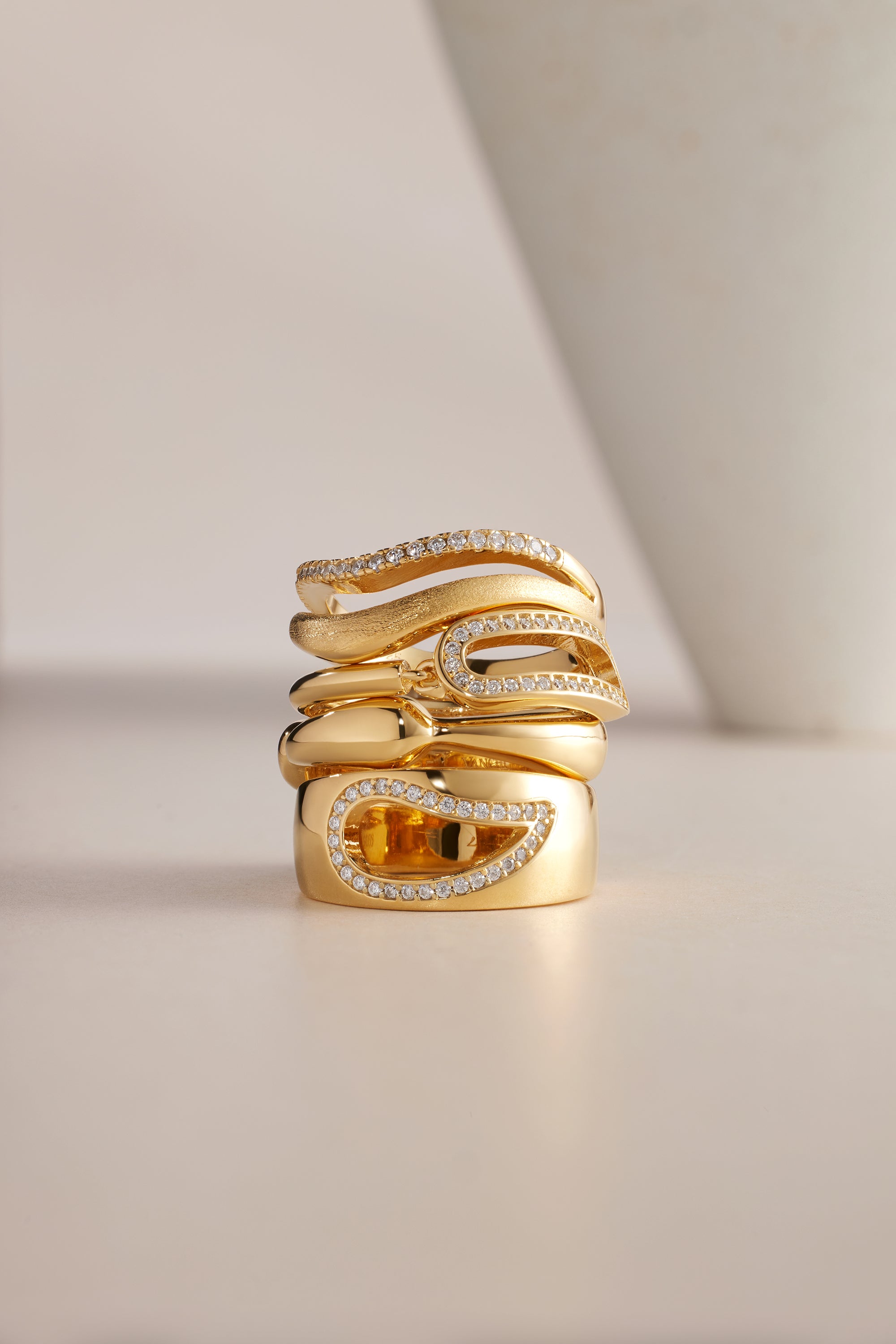Three elegant gold rings, including the Colette Link Droop Ring in Gold with unique curves, are stacked together. The rings feature sparkling diamonds and one has a rectangular yellow gemstone. A blurred white object appears in the background.