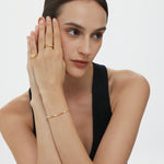 A woman with dark hair in a low bun wears a sleeveless black top and the Noor Pavé Triple Band Ring in Gold, resting her head on her hands and gazing thoughtfully to the side against a plain light background.