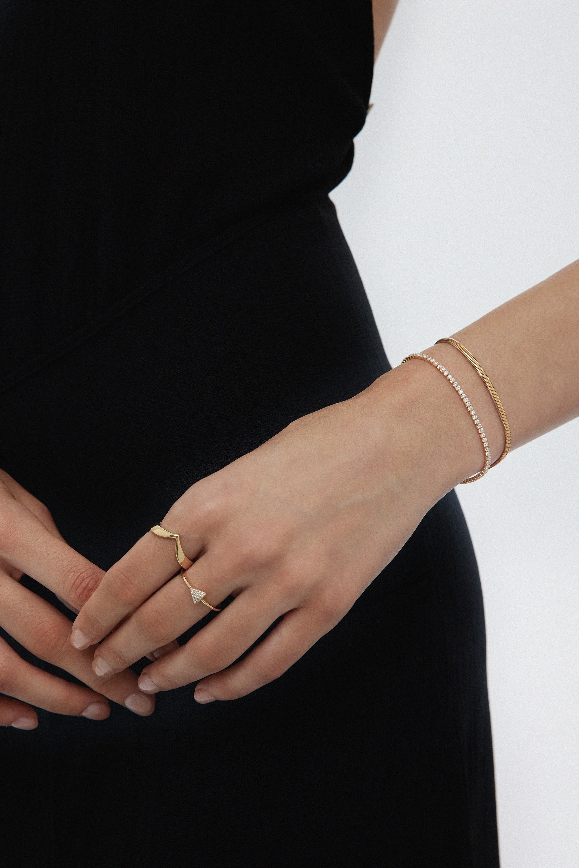 A person in black showcases their left hand with the Veda Plain Chevron Ring in Gold, plus two gold bracelets, against a plain white background.