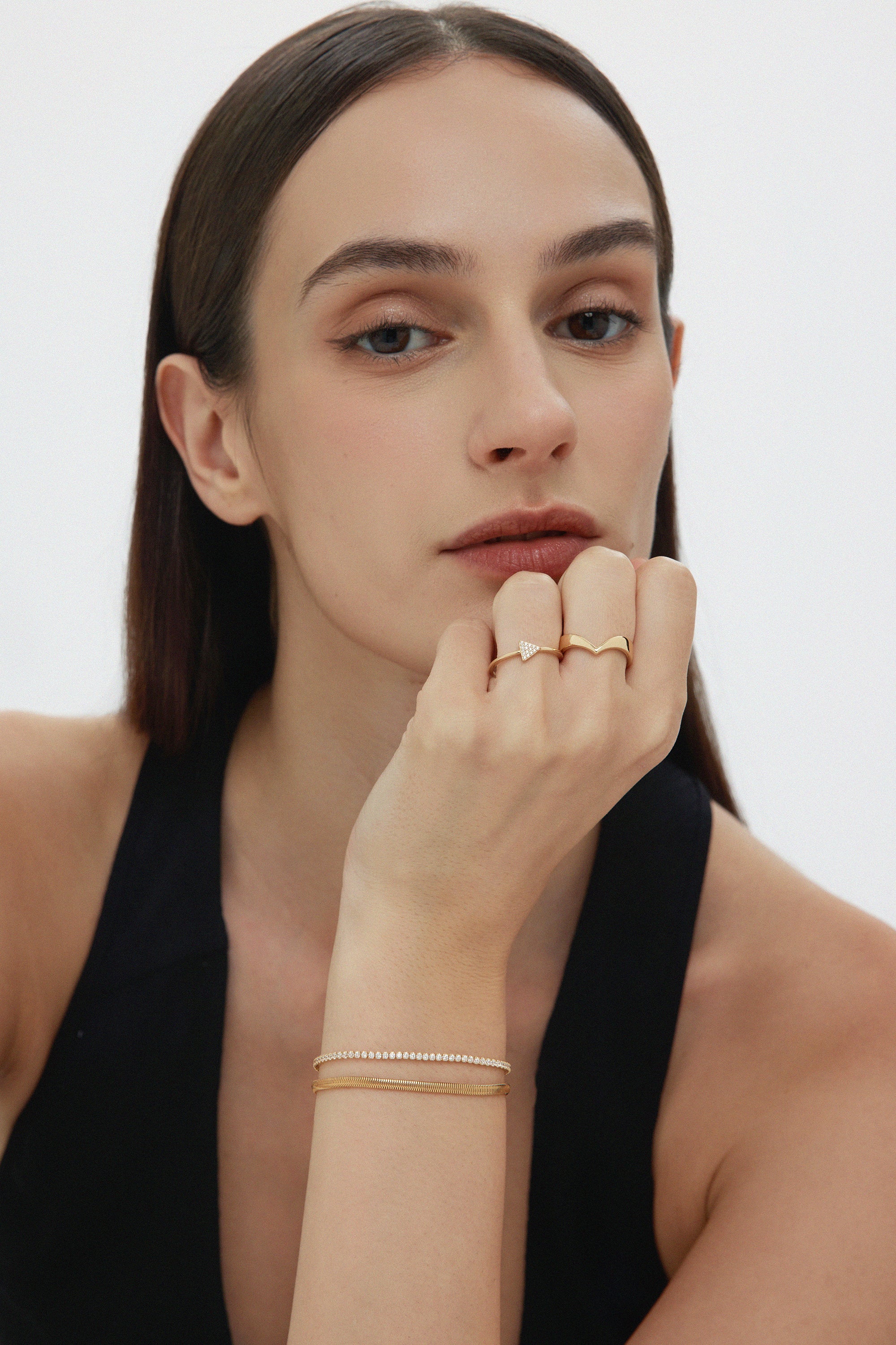 A woman with straight brown hair and natural makeup rests her chin on her hand, showcasing the Veda Plain Chevron Ring in Gold. She wears a sleeveless black top and looks directly at the camera.
