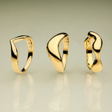 The Vava Plain Chunky Curved Wave Ring in Gold, with its modern sculptural form, stands upright on a reflective surface against a green-to-beige gradient background—a striking showcase of minimalist jewelry.