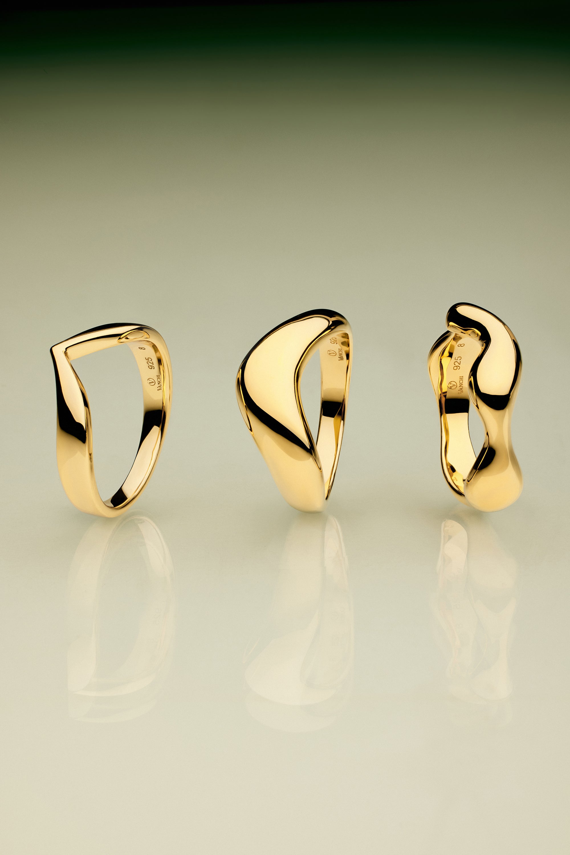 The Vava Plain Chunky Curved Wave Ring in Gold, with its modern sculptural form, stands upright on a reflective surface against a green-to-beige gradient background—a striking showcase of minimalist jewelry.
