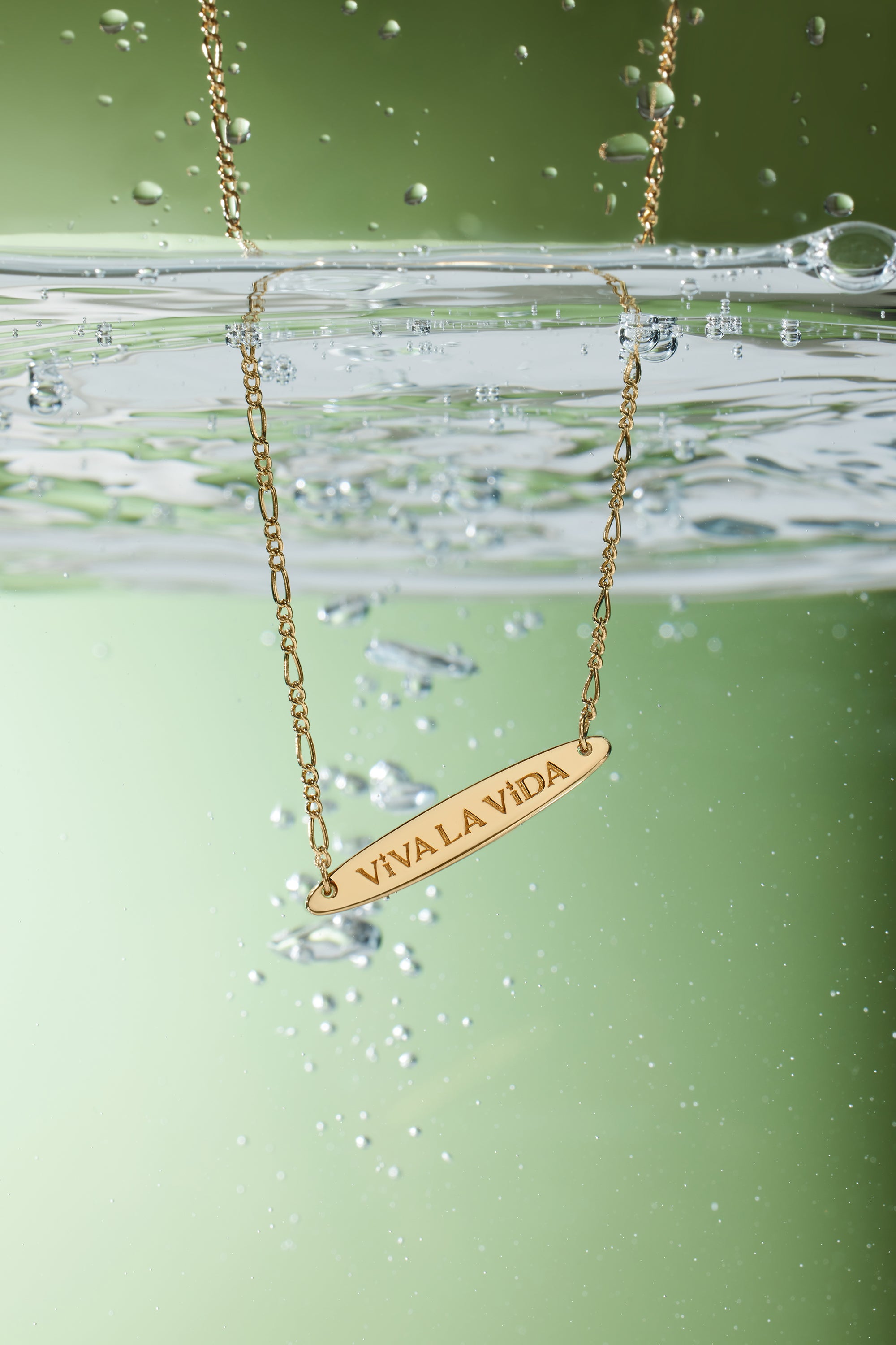 The Viva La Vida Bar Chain Necklace in Gold features VIVA LA VIDA engraved on a partially submerged pendant with bubbles and an adjustable figaro chain, set against a green background.