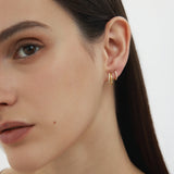 A close-up of a woman with straight dark hair wearing two Kiki Crystal Huggie Earrings in Gold, perfect for ear stacking, in her left ear against a plain light background. Only the left side of her face is visible.