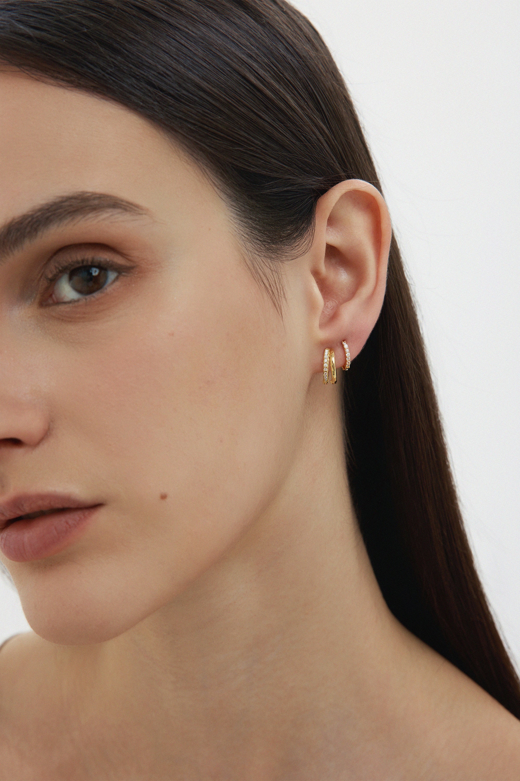A close-up of a woman with straight dark hair wearing two Kiki Crystal Huggie Earrings in Gold, perfect for ear stacking, in her left ear against a plain light background. Only the left side of her face is visible.
