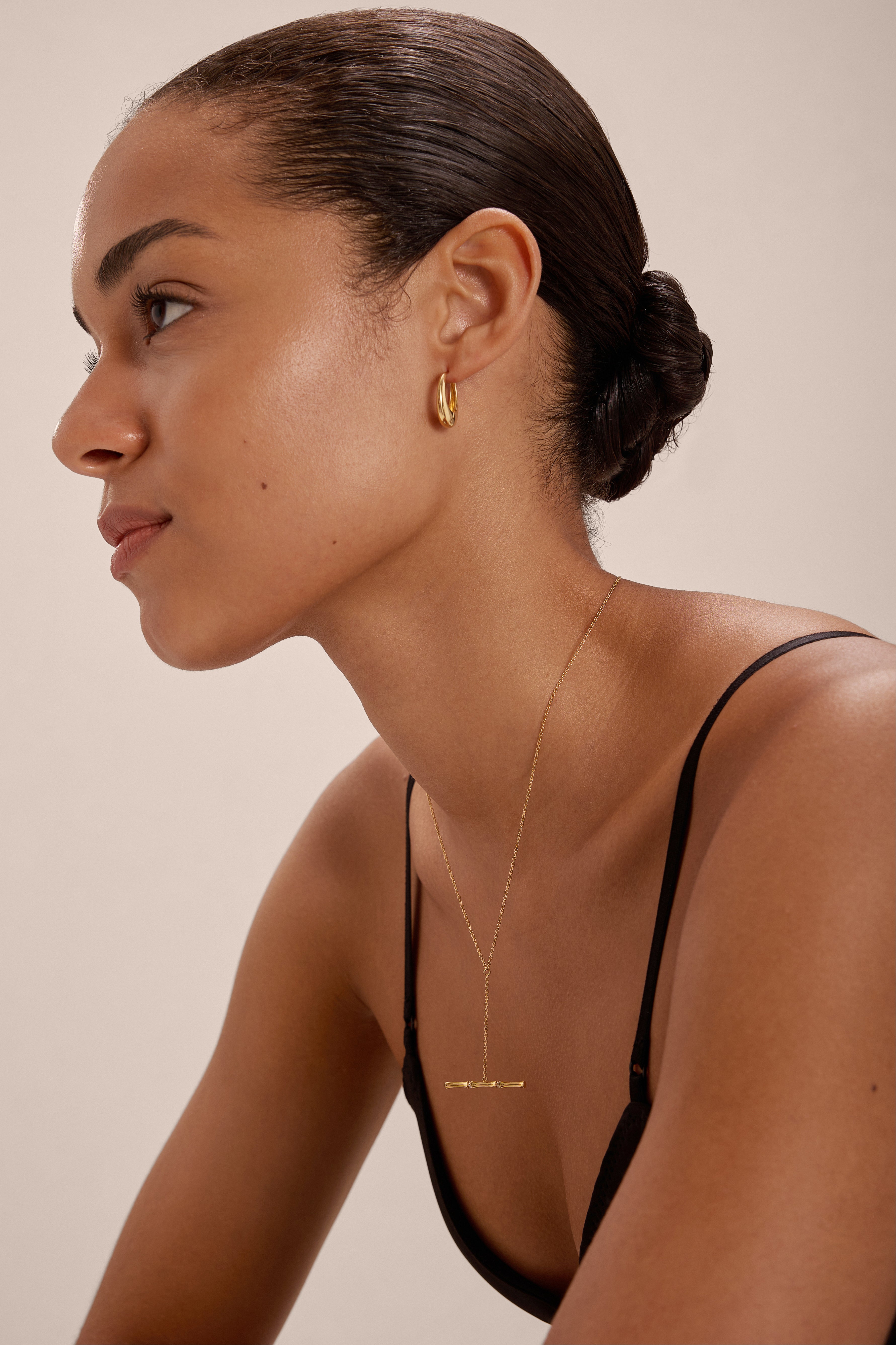 A woman with smooth, medium-brown skin and a low bun wears a black top, the Lila Medium Round Dome Hoop Earrings in Gold, and a delicate gold necklace. She faces left against a softly lit, neutral background.