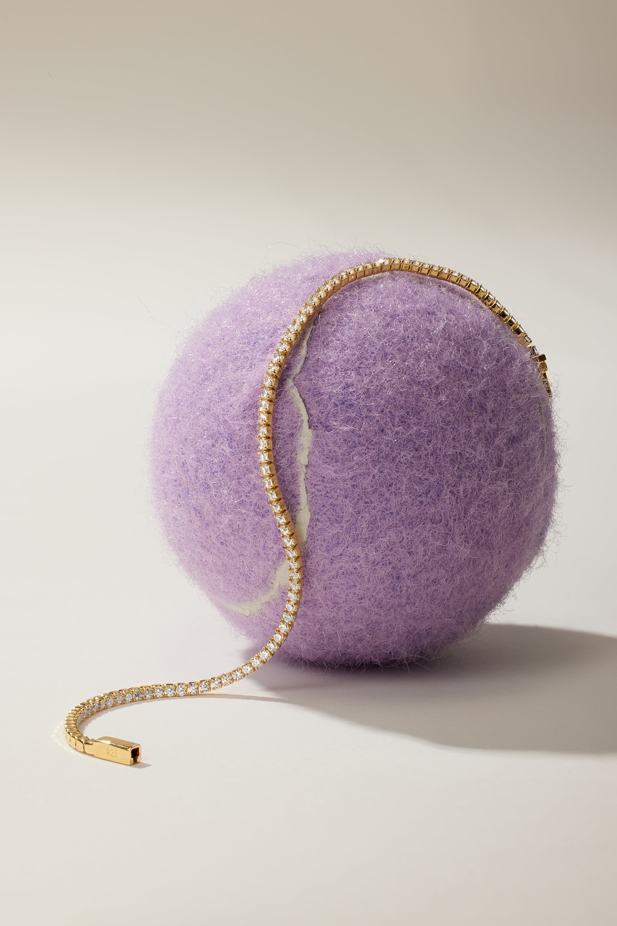 The Lumi Tennis Bracelet in Gold, adorned with sparkling cubic zirconia gemstones, is elegantly draped over a large fuzzy purple ball against a simple light background.