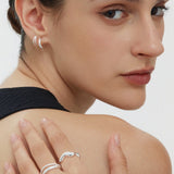 A woman with slicked-back hair wears Mae Twist Double Huggie Earrings in Silver and silver rings, resting her hand on her bare shoulder as she looks over her shoulder at the camera.