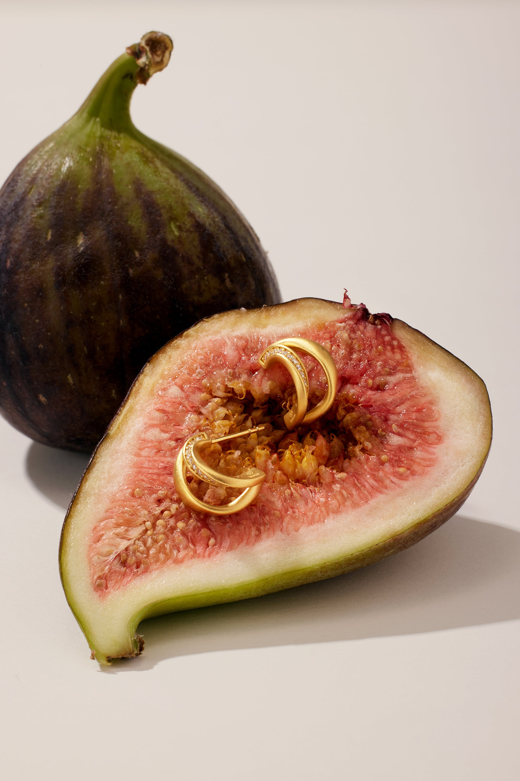 A halved fig with pink flesh holds two Nadine Pavé Twisted Stud Earrings in Gold, while a whole fig stands in the background, both illuminated by soft, natural light.