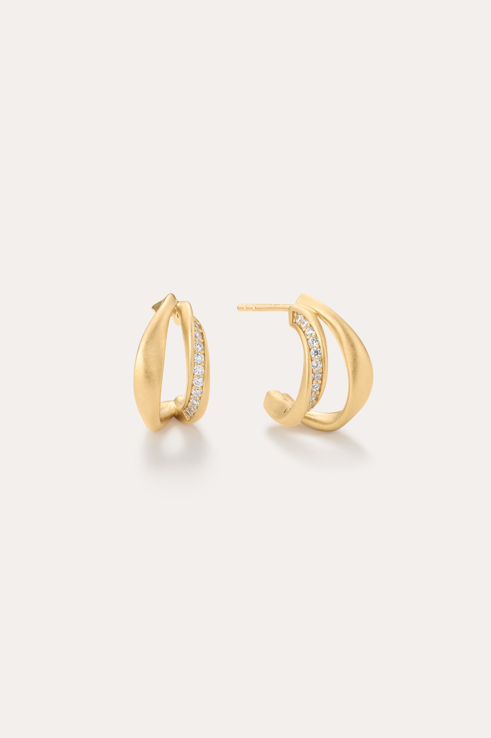 Nadine Pavé Twisted Stud Earrings in Gold feature a modern, sculptural design with a row of sparkling gemstones set along the inner edge and are displayed against a light neutral background.