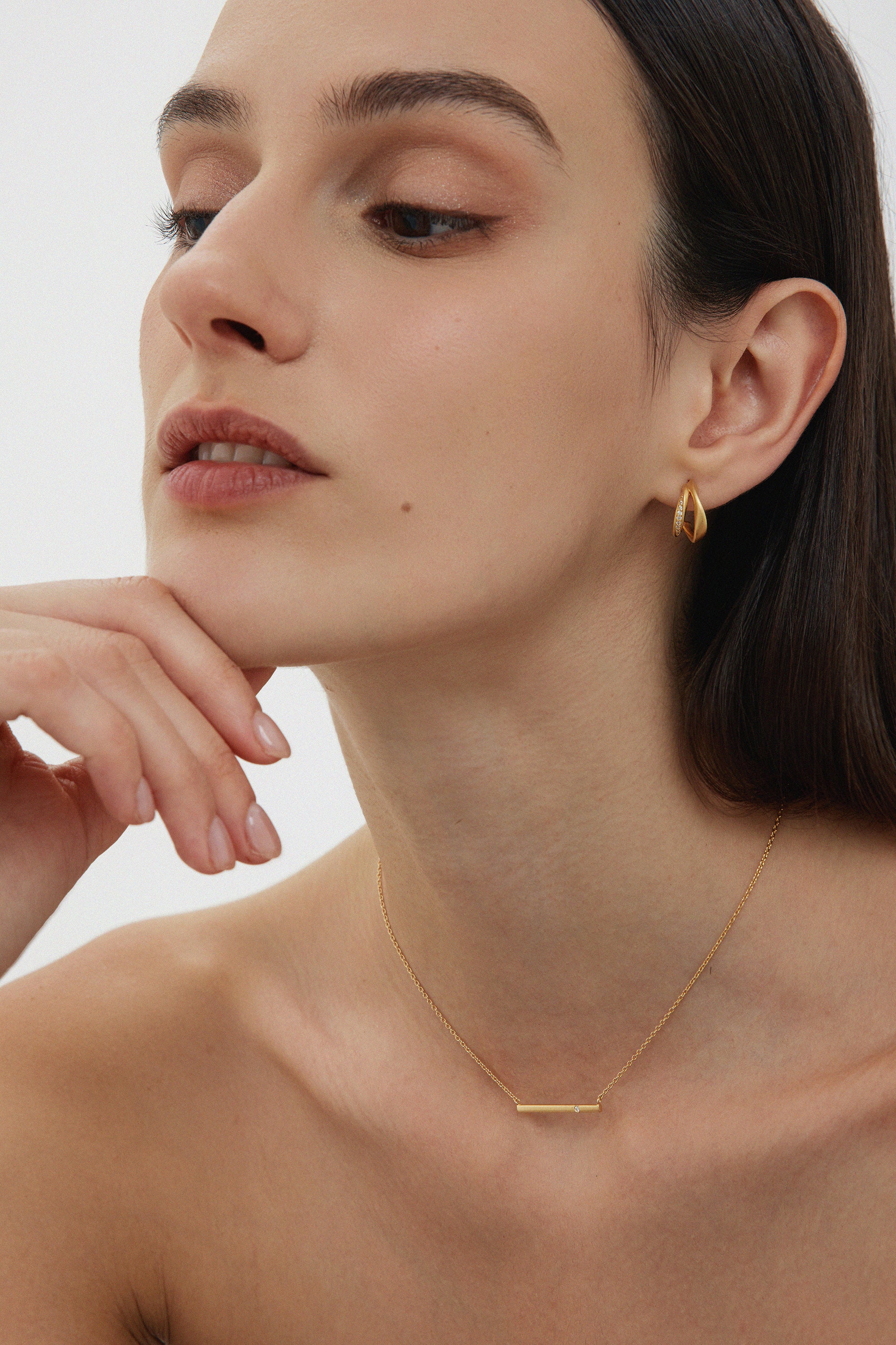 A woman with straight dark hair poses thoughtfully, resting her chin on her hand. She wears Nadine Pavé Twisted Stud Earrings in Gold and a delicate gold bar necklace, with minimal makeup against a plain, light background.