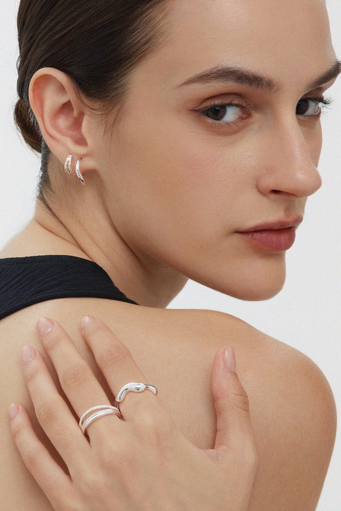 A woman with slicked-back hair wears a sleeveless black top, silver hoop earrings, and the Noor Pavé Triple Band Ring in Silver as she looks over her shoulder at the camera against a plain white background.