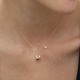 A close-up of a woman’s neck wearing the Nora Double Pendant Necklace in Gold, featuring a small round sculptural pendant and a tiny dangling cubic zirconia, set against light skin and a black top.
