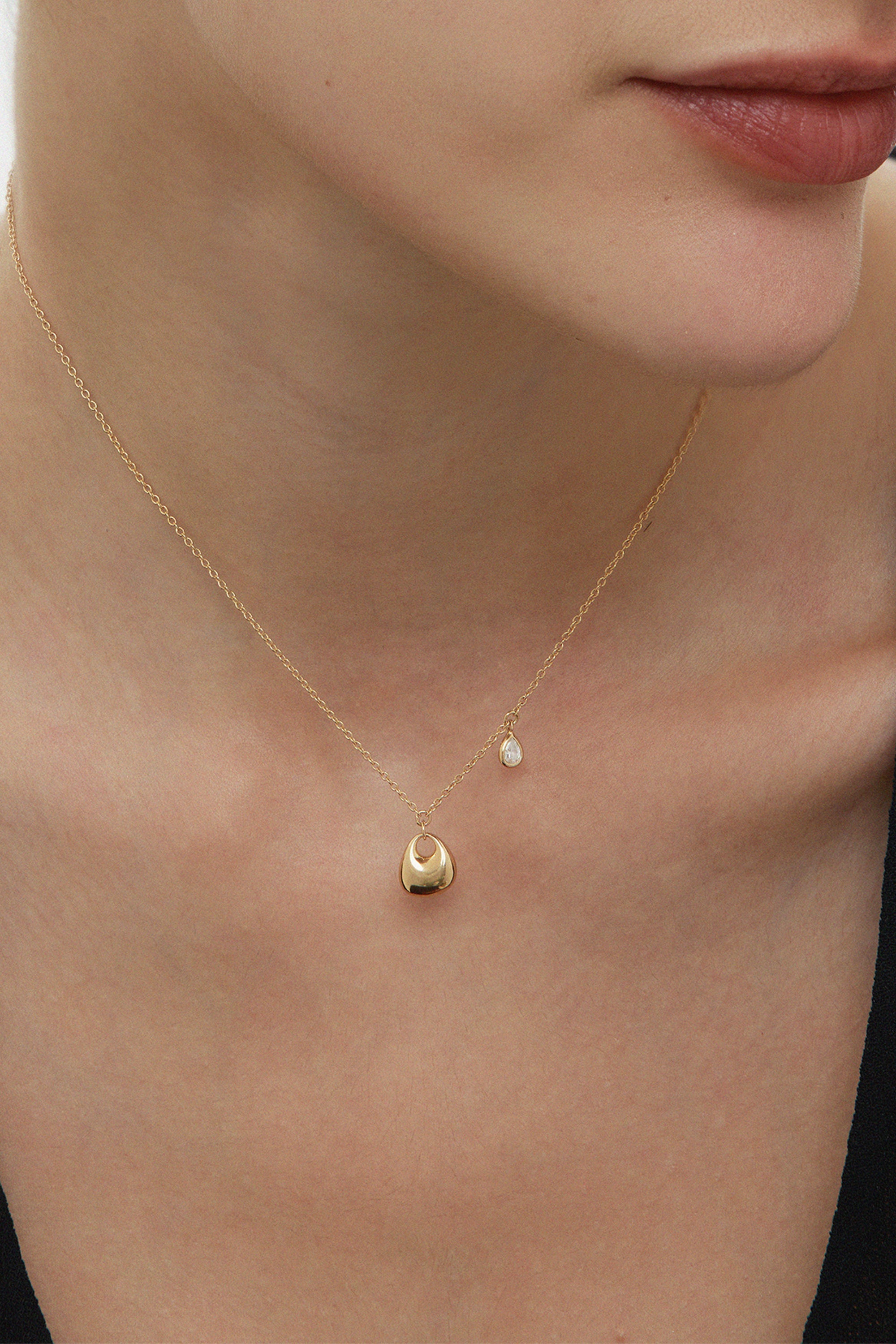 A close-up of a woman’s neck wearing the Nora Double Pendant Necklace in Gold, featuring a small round sculptural pendant and a tiny dangling cubic zirconia, set against light skin and a black top.