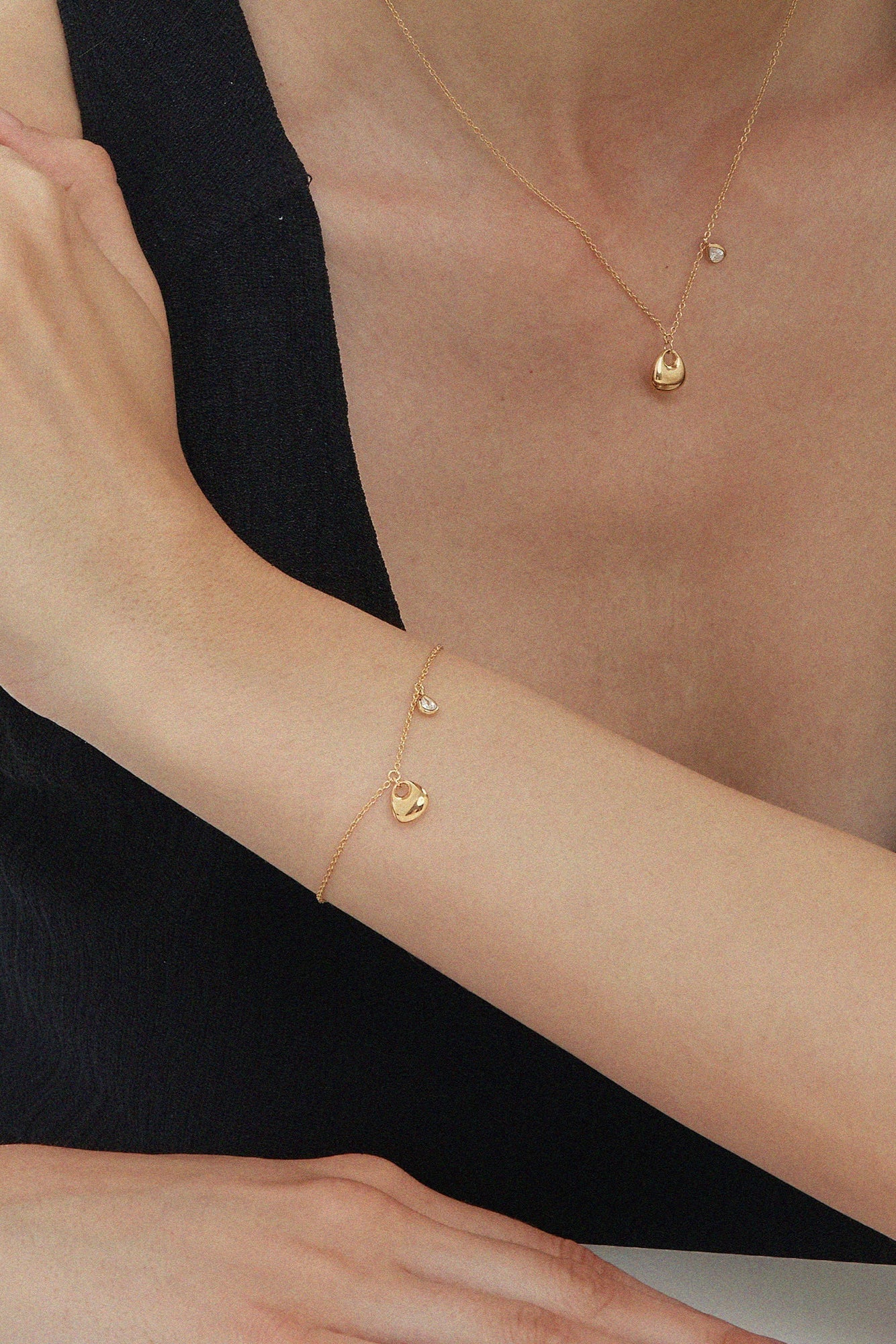A person in a sleeveless black top rests their arm on a table, showcasing the Nora Double Pendant Bracelet in Gold alongside other gold jewelry, with their hand touching their upper arm against a plain white background.