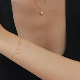 A woman in a sleeveless black top rests her arm on a surface, wearing delicate gold jewelry, including the Nora Double Pendant Necklace in Gold, plus a matching bracelet and ring. Her other hand gently touches her shoulder against a white background.